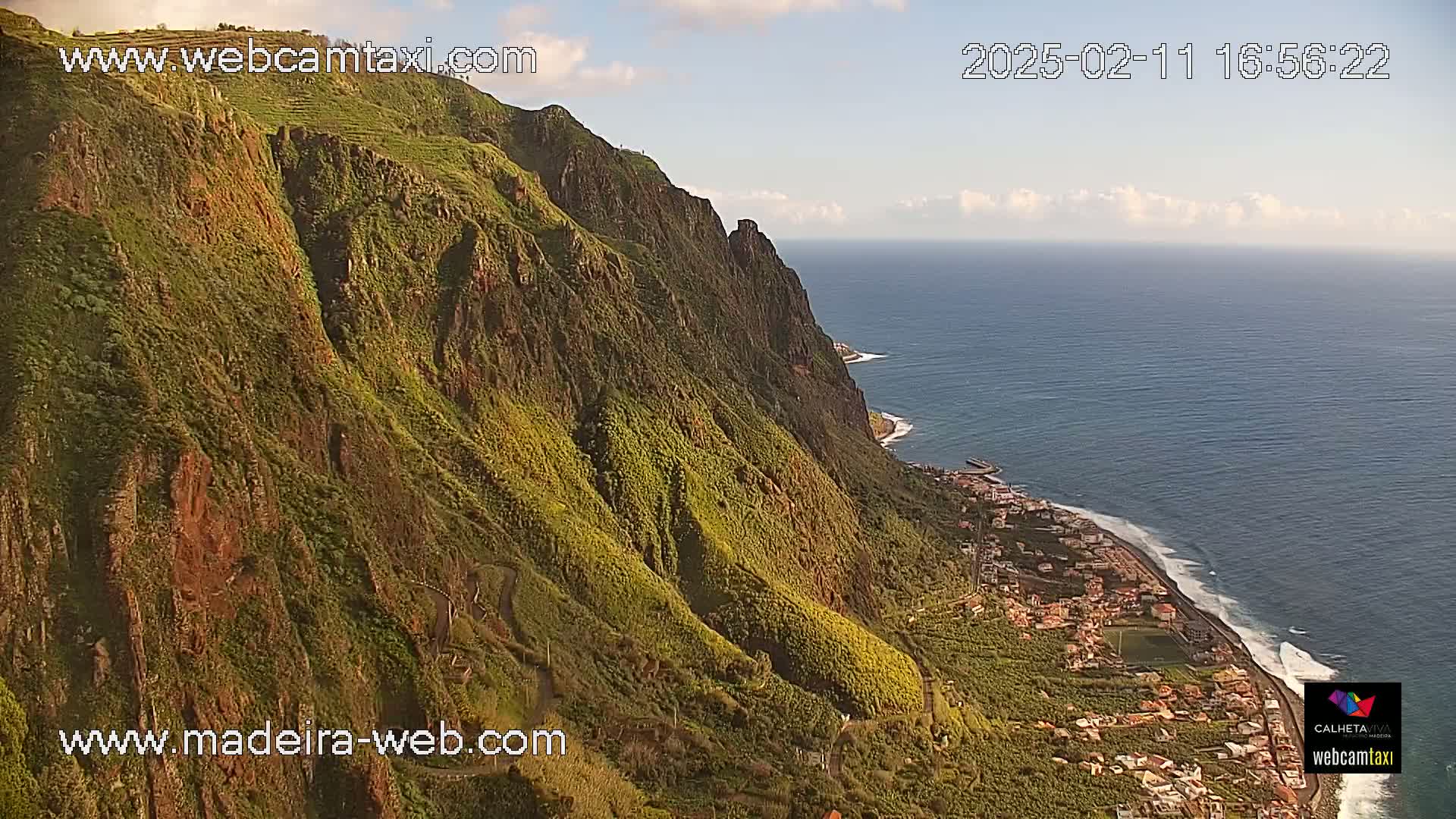 A steep, green cliffside overlooks a coastline with a town and ocean under partly cloudy skies.