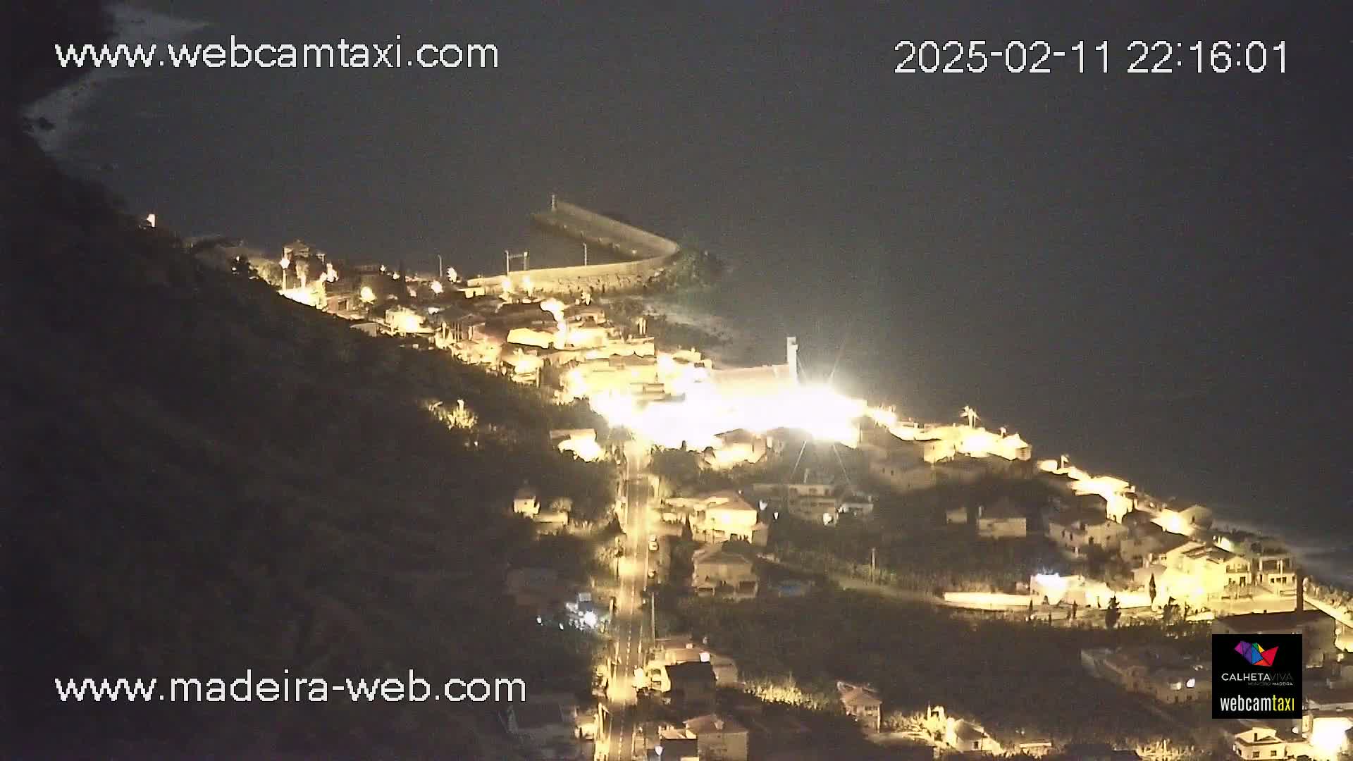 A nighttime aerial view shows a coastal town illuminated on a hillside overlooking a dark body of water.