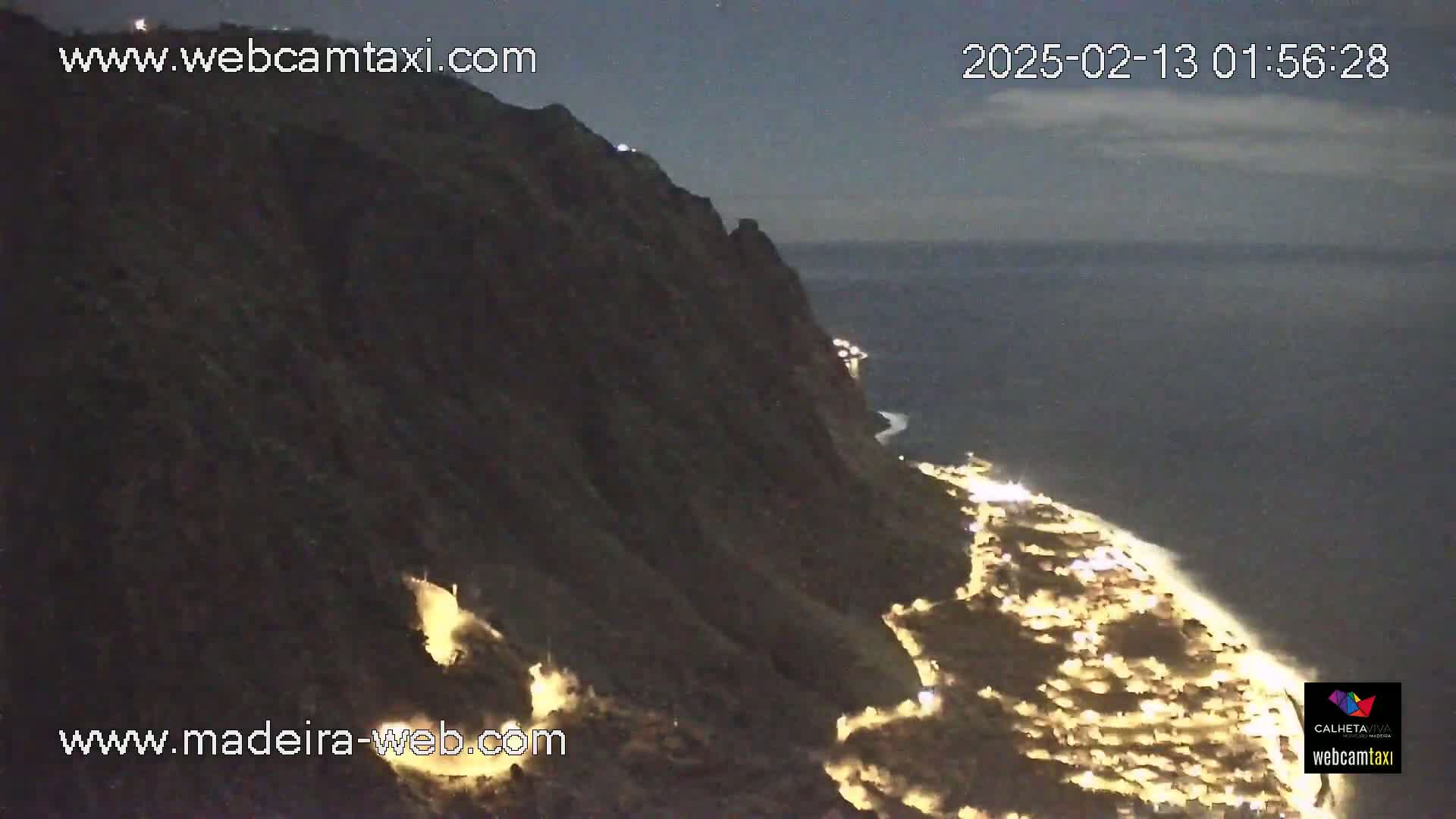 A nighttime view shows a dark cliffside overlooking a coastal town illuminated by many lights, under a mostly clear night sky.