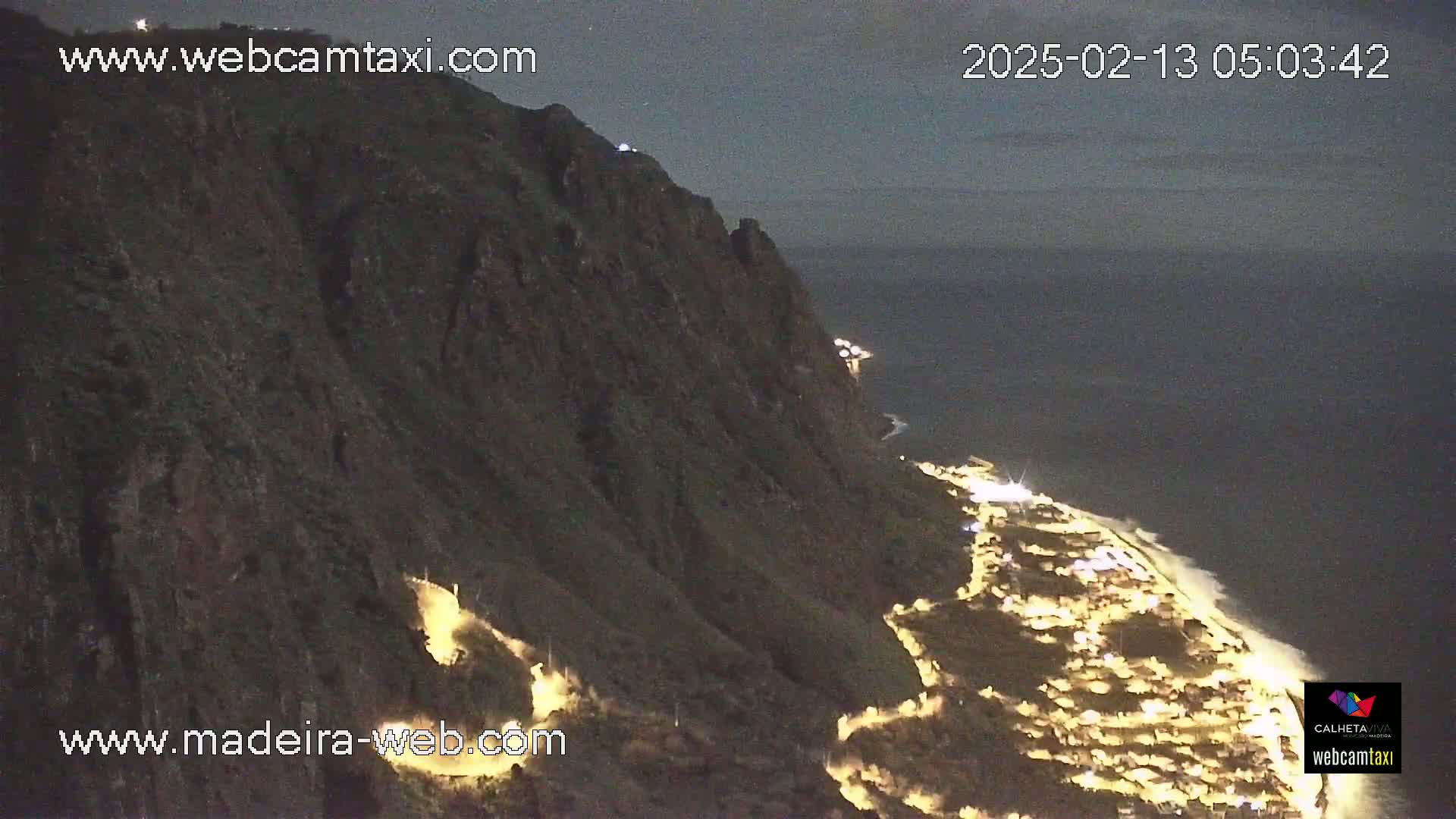 A nighttime, coastal view shows a dark cliff face overlooking a town illuminated by lights along the shoreline and the ocean under a dark sky.