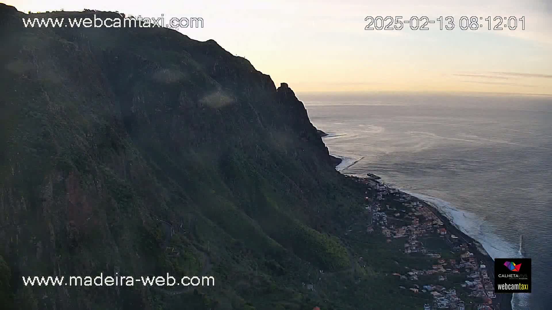 A high-angle, long shot shows a steep, dark green cliff overlooking a coastal town and the ocean at dawn under a pale, clear sky.