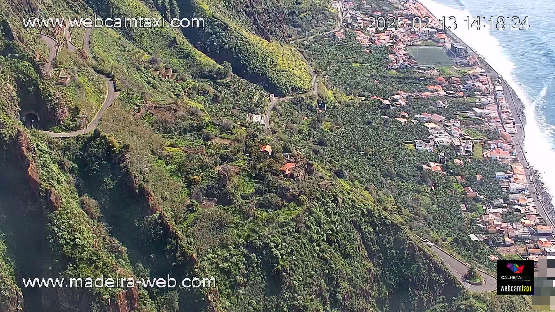 A winding road curves through lush green hillsides down to a coastal town with ocean waves visible in the distance on a sunny day.