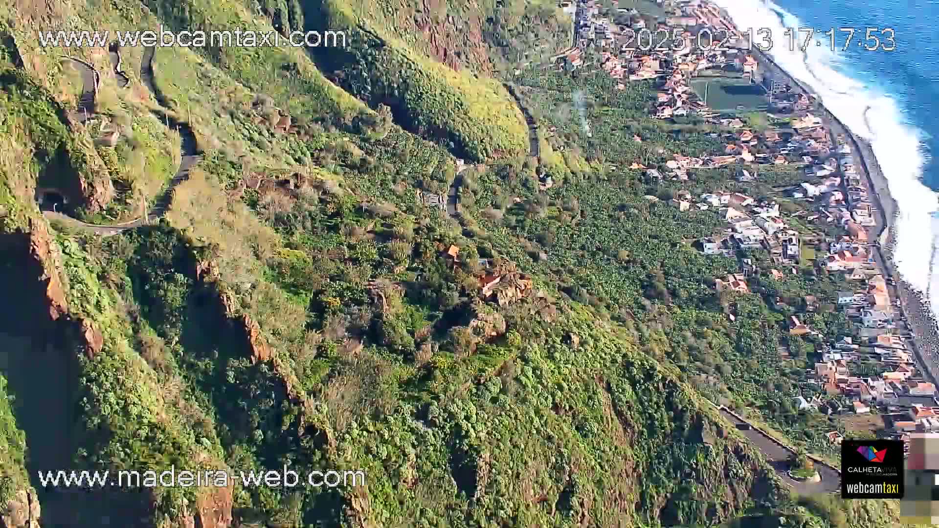 A high-angle view shows a winding road descending a lush, green, rocky hillside toward a coastal town and ocean with breaking waves under a sunny sky.