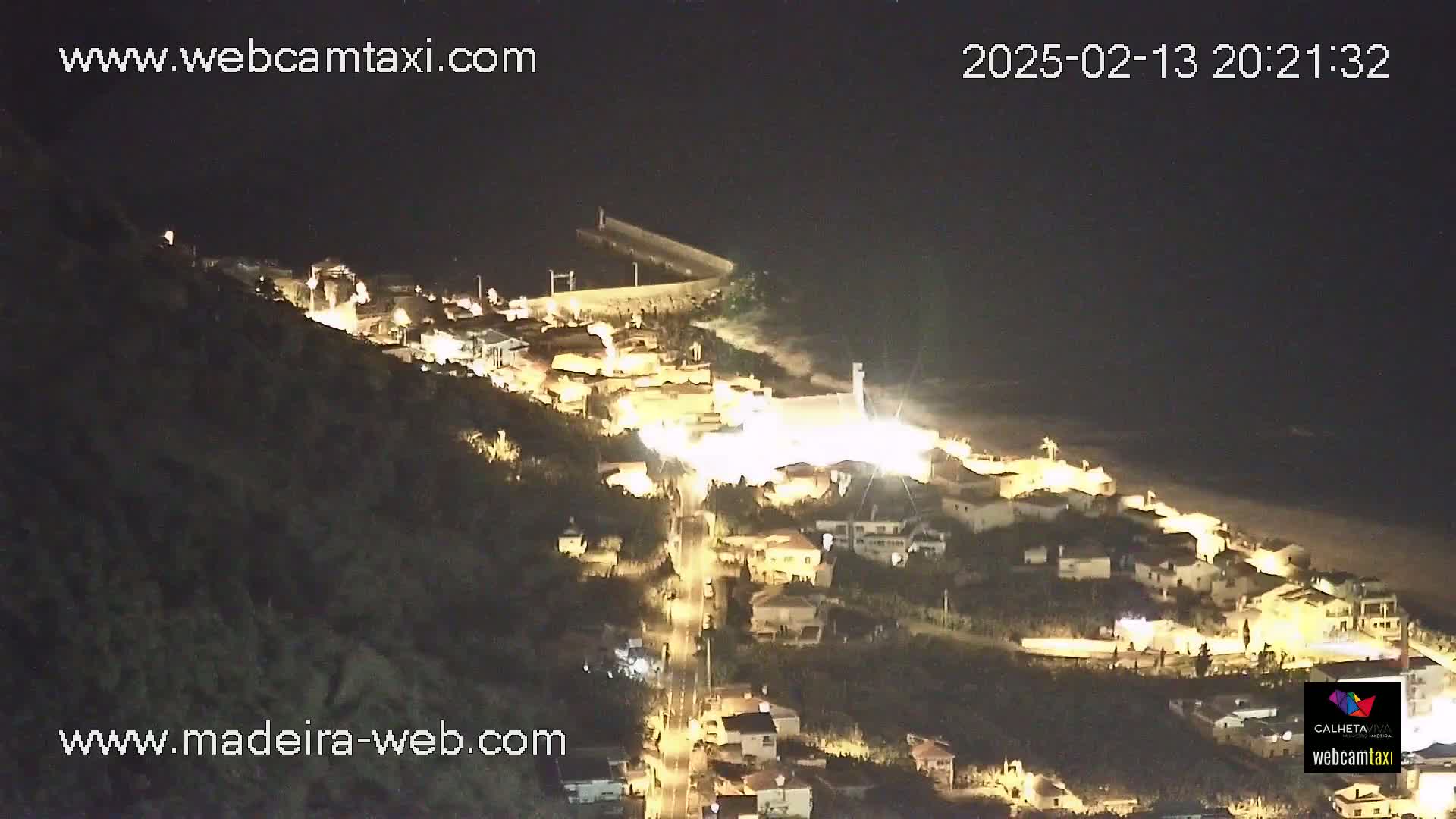 A nighttime, aerial view shows a coastal town illuminated with lights, situated on a hillside next to a dark ocean with a breakwater.