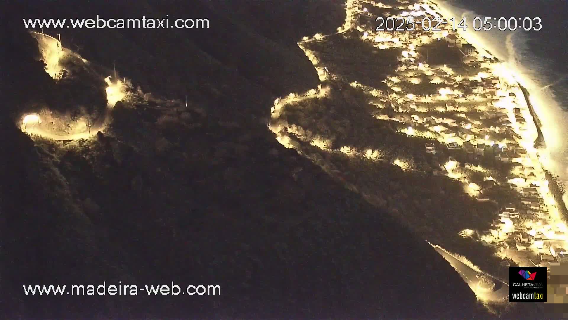 A nighttime aerial view shows a coastal town illuminated by streetlights, situated on a hillside next to a dark ocean.