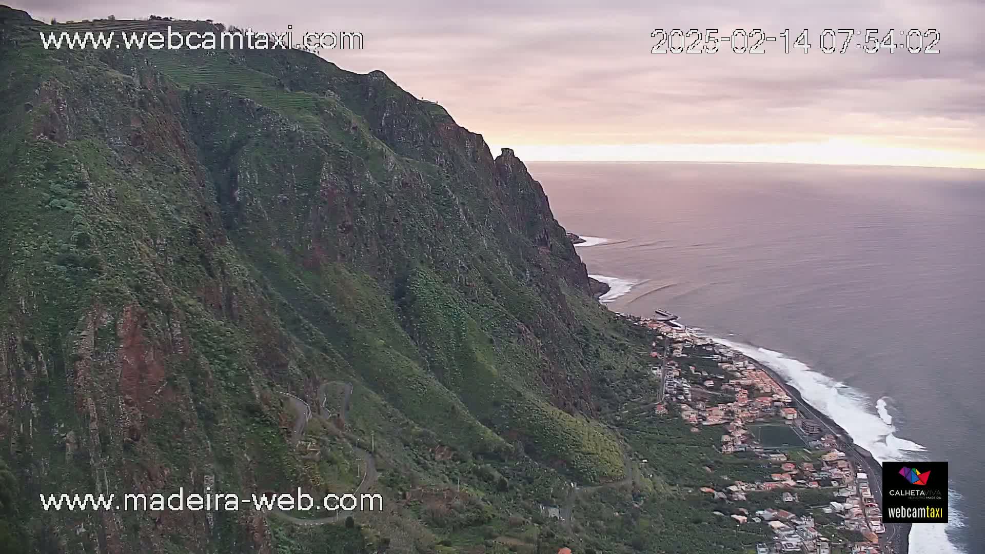 A steep, green cliffside overlooks a coastal town and ocean under a partly cloudy, pinkish sky at dawn or dusk.
