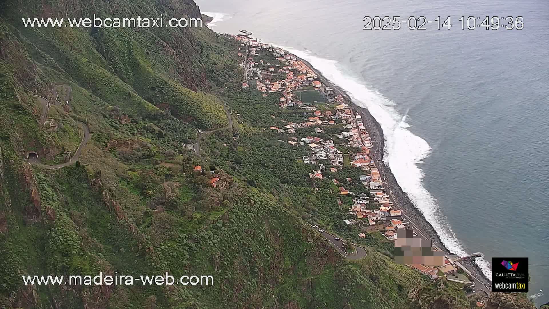An aerial view shows a coastal town nestled between a steep, green mountain and a dark-blue ocean with white-capped waves under a cloudy sky.