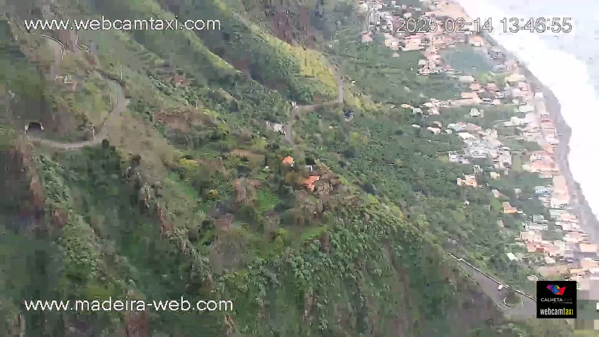 A winding road snakes down a lush, green mountainside toward a coastal village, with the ocean visible in the background on an overcast day.