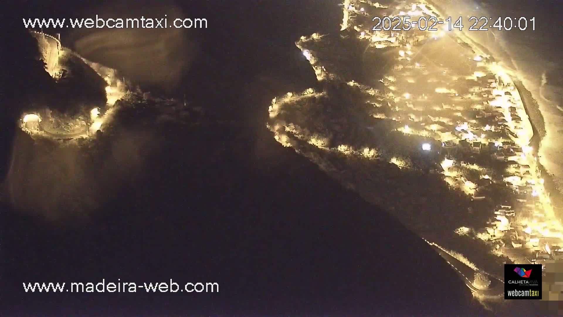 A nighttime aerial view shows a coastal town illuminated by streetlights, with a winding road leading up a dark hillside.
