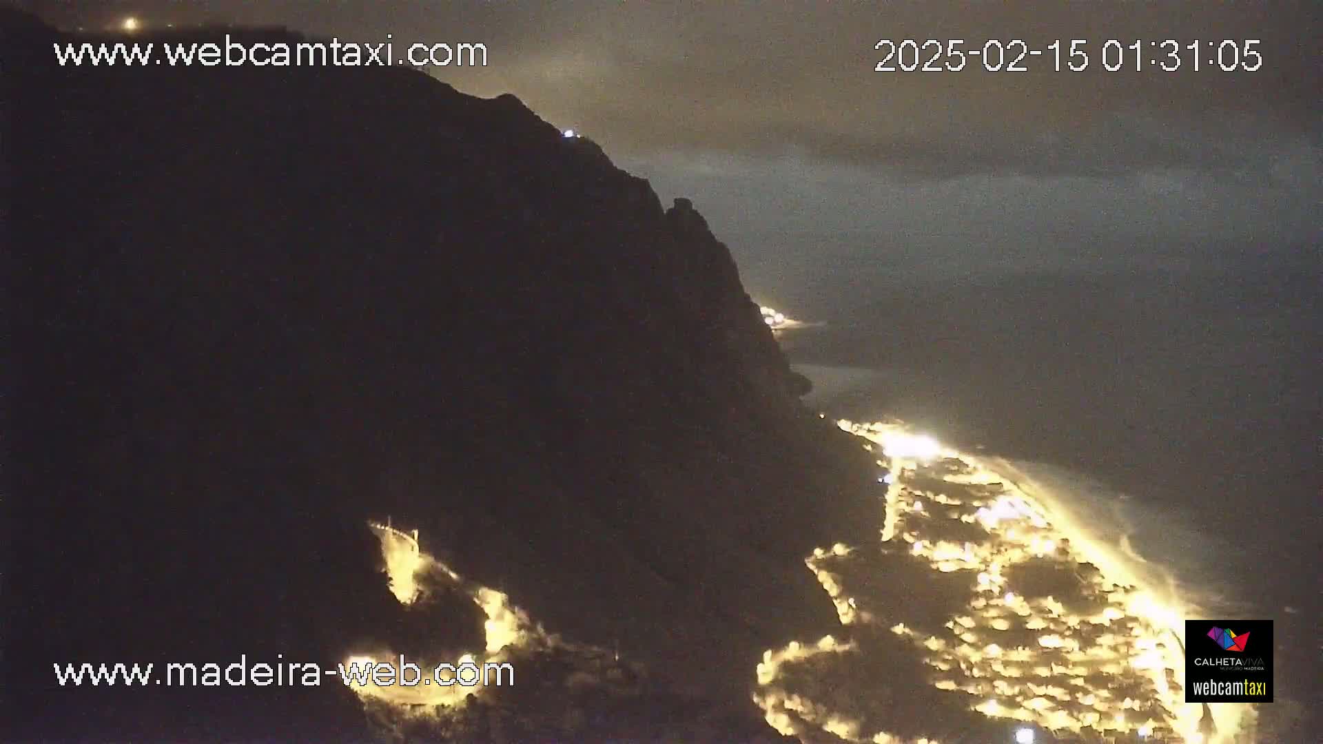 A nighttime coastal view shows a dark cliff overlooking a town illuminated by lights along the shoreline under a cloudy sky.