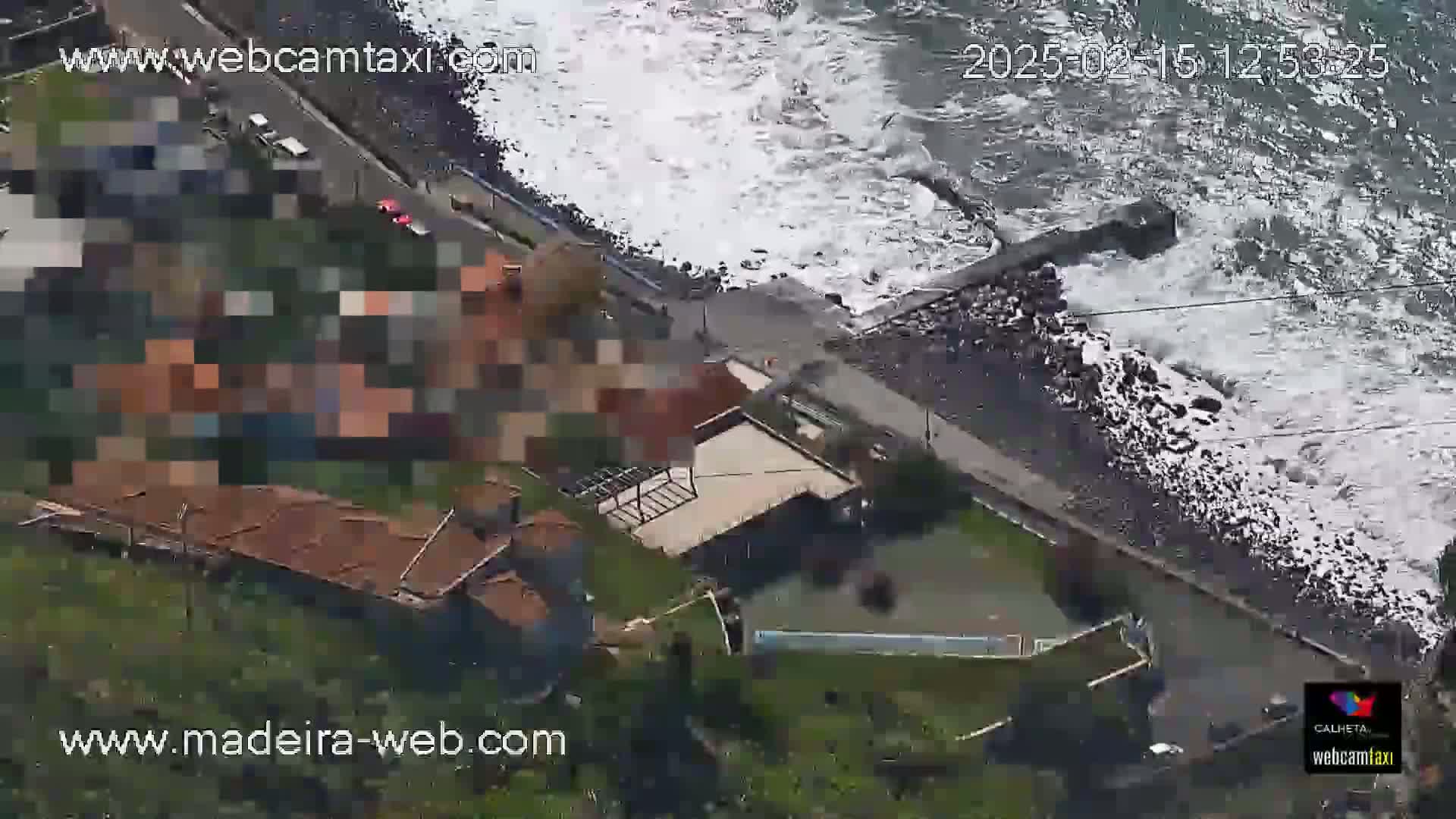 An aerial view shows a rocky coastline with crashing waves, a paved walkway, and buildings along a hillside.