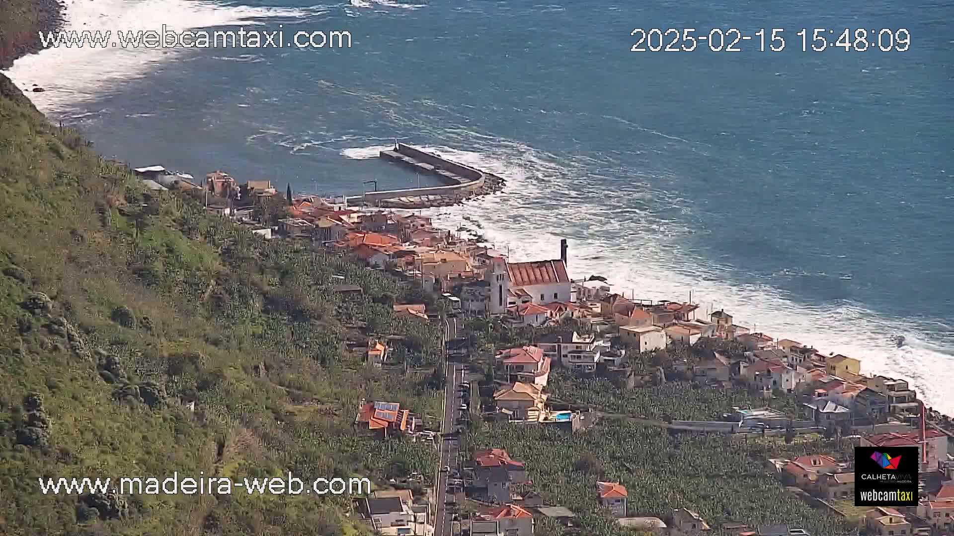 A coastal town with a church nestled between a hillside and a rocky shoreline with moderate waves on a sunny day.