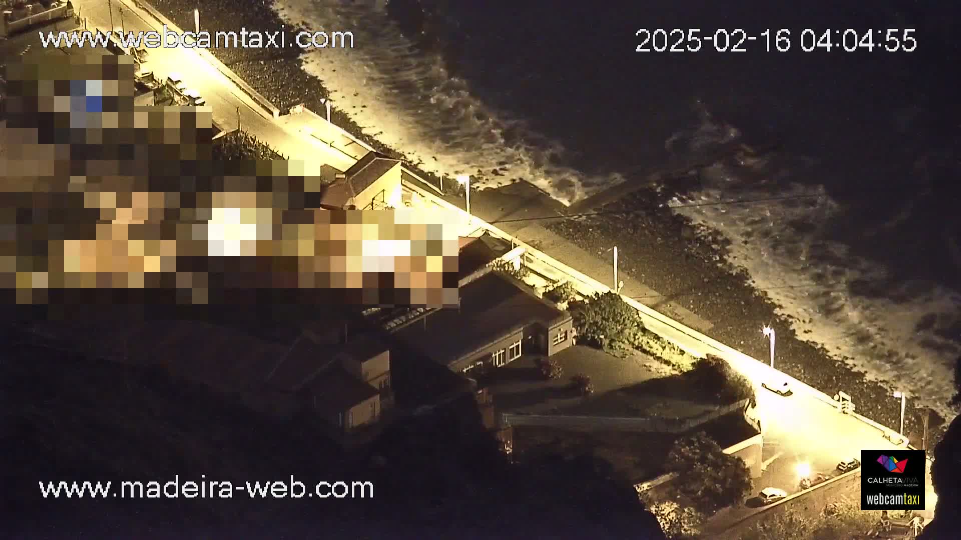 A nighttime aerial view shows a coastal road with buildings and a few vehicles, next to a rocky shoreline with waves crashing under a dark sky.