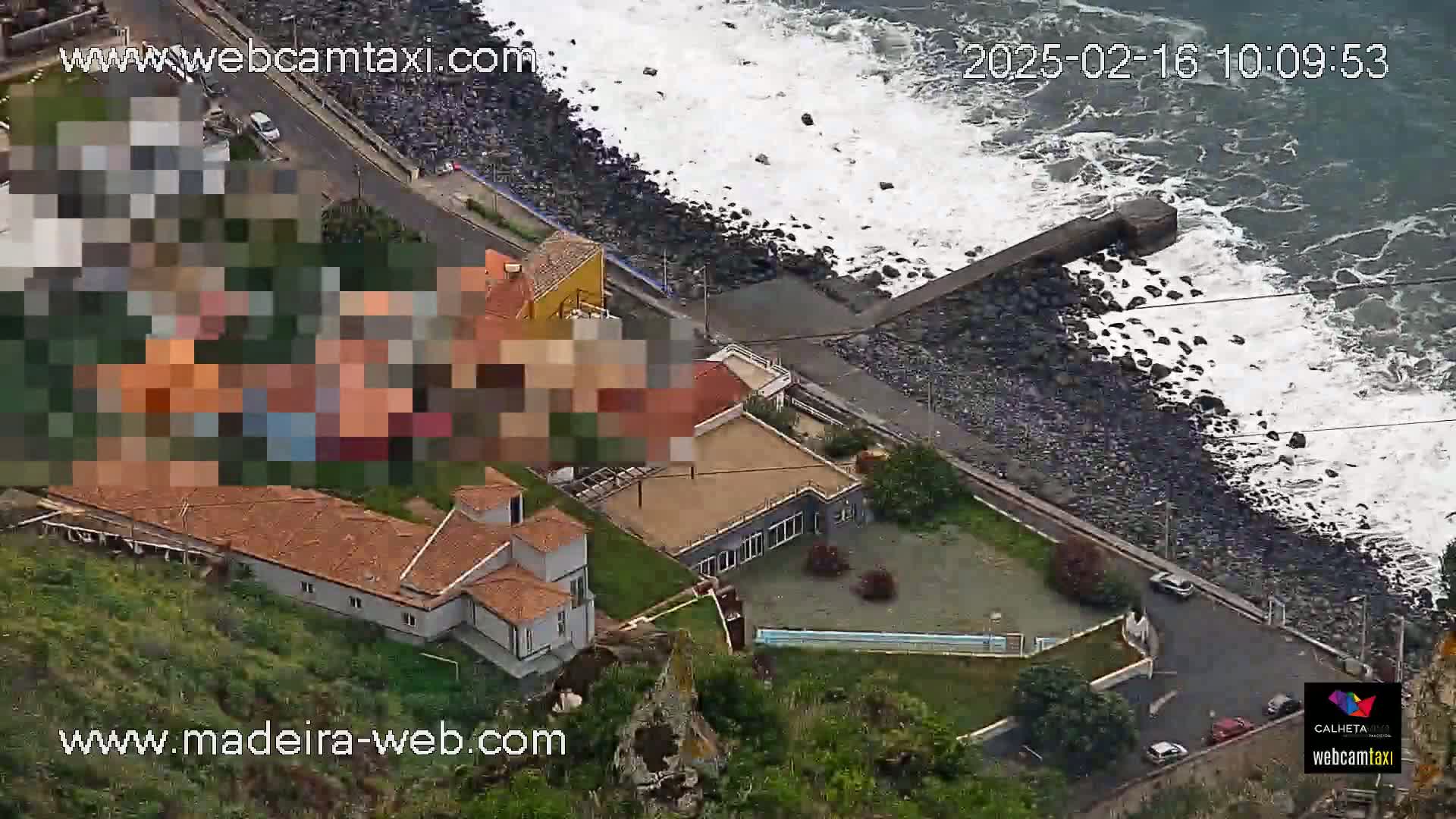 An aerial view shows a rocky coastline with crashing waves, a road with cars, and several buildings including a large house with a swimming pool, all under partly cloudy skies.
