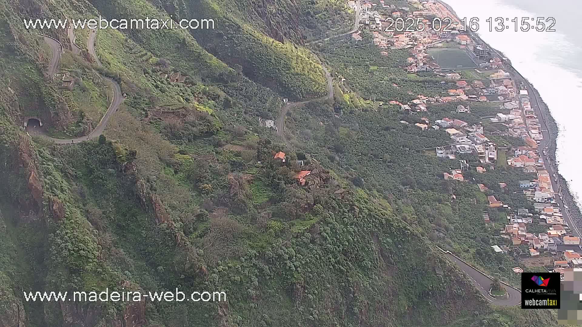 A winding road snakes down a lush, green hillside toward a coastal town and ocean, under a mostly clear sky.