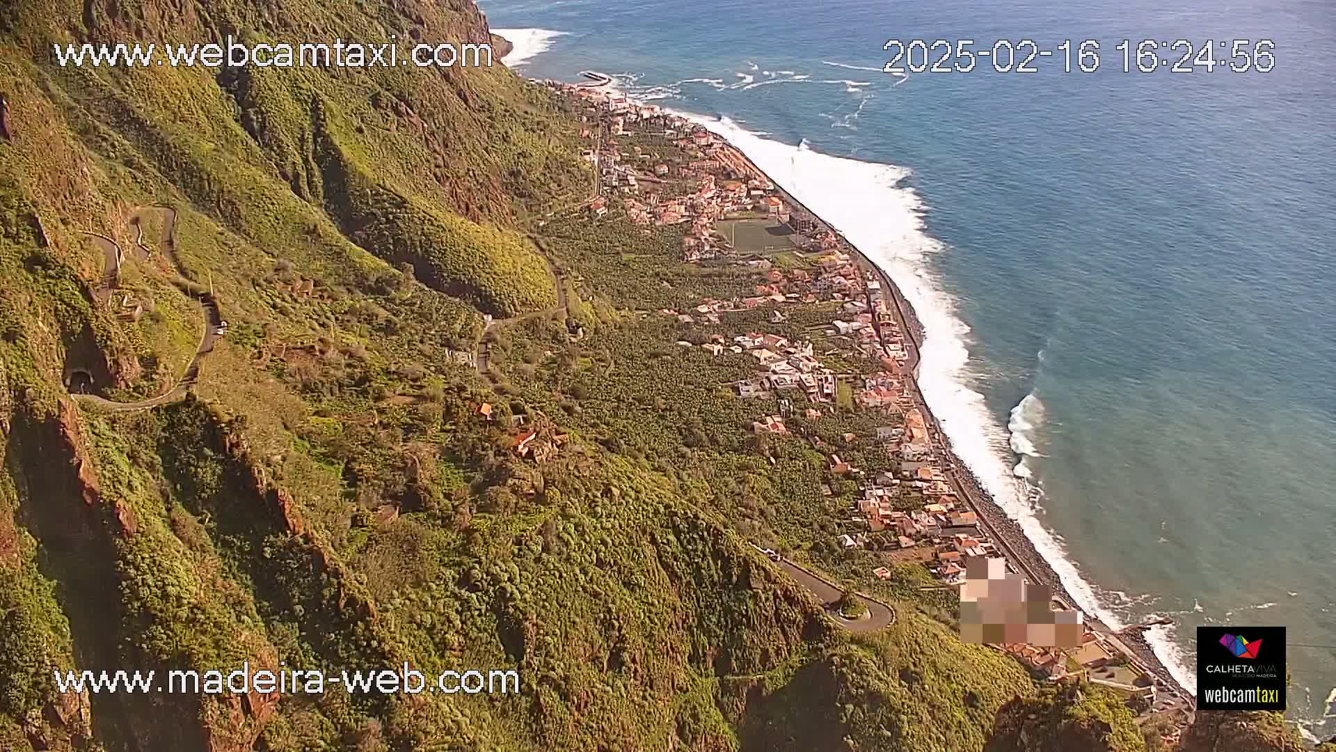 A high-angle view shows a winding road along a steep, green hillside overlooking a coastline with breaking waves on a sunny day.