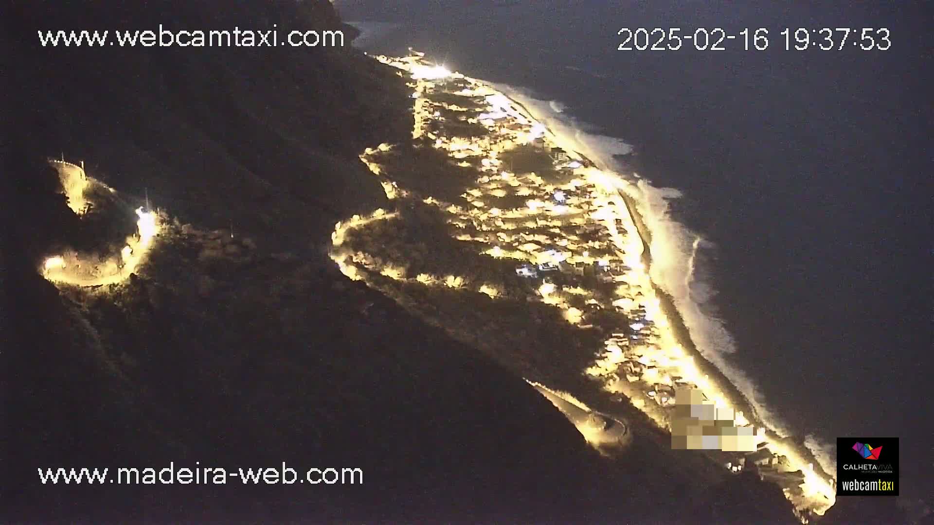 A nighttime aerial view shows a coastal town illuminated along a dark, wavy ocean.