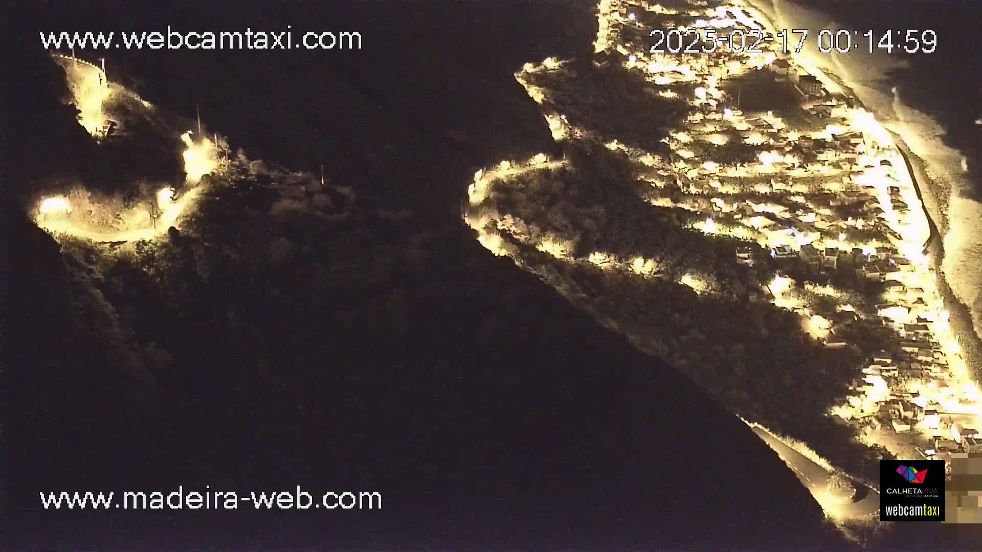 An aerial nighttime view shows a coastal town illuminated, nestled between dark hillsides with winding roads visible.