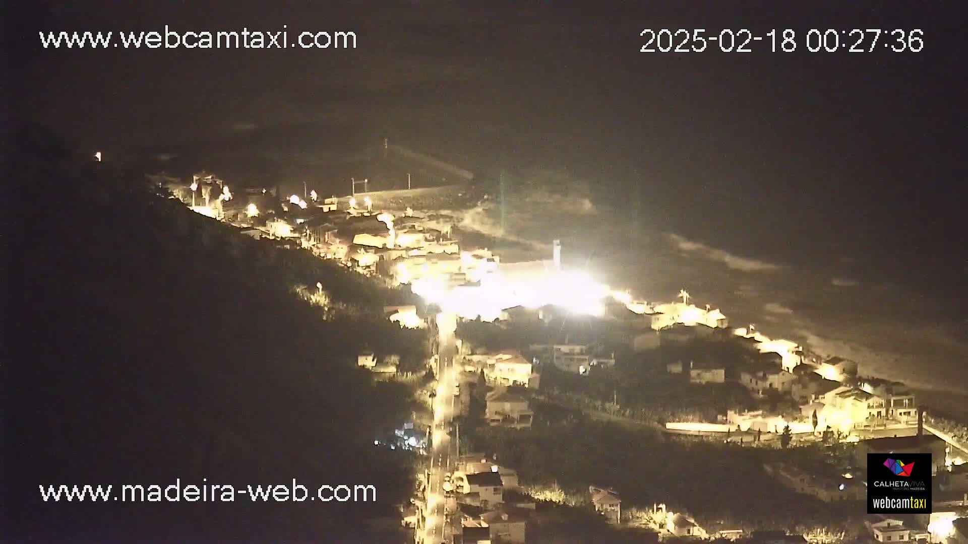 A nighttime, high-angle view of a coastal town illuminated by streetlights, with dark, wavy ocean visible in the background.