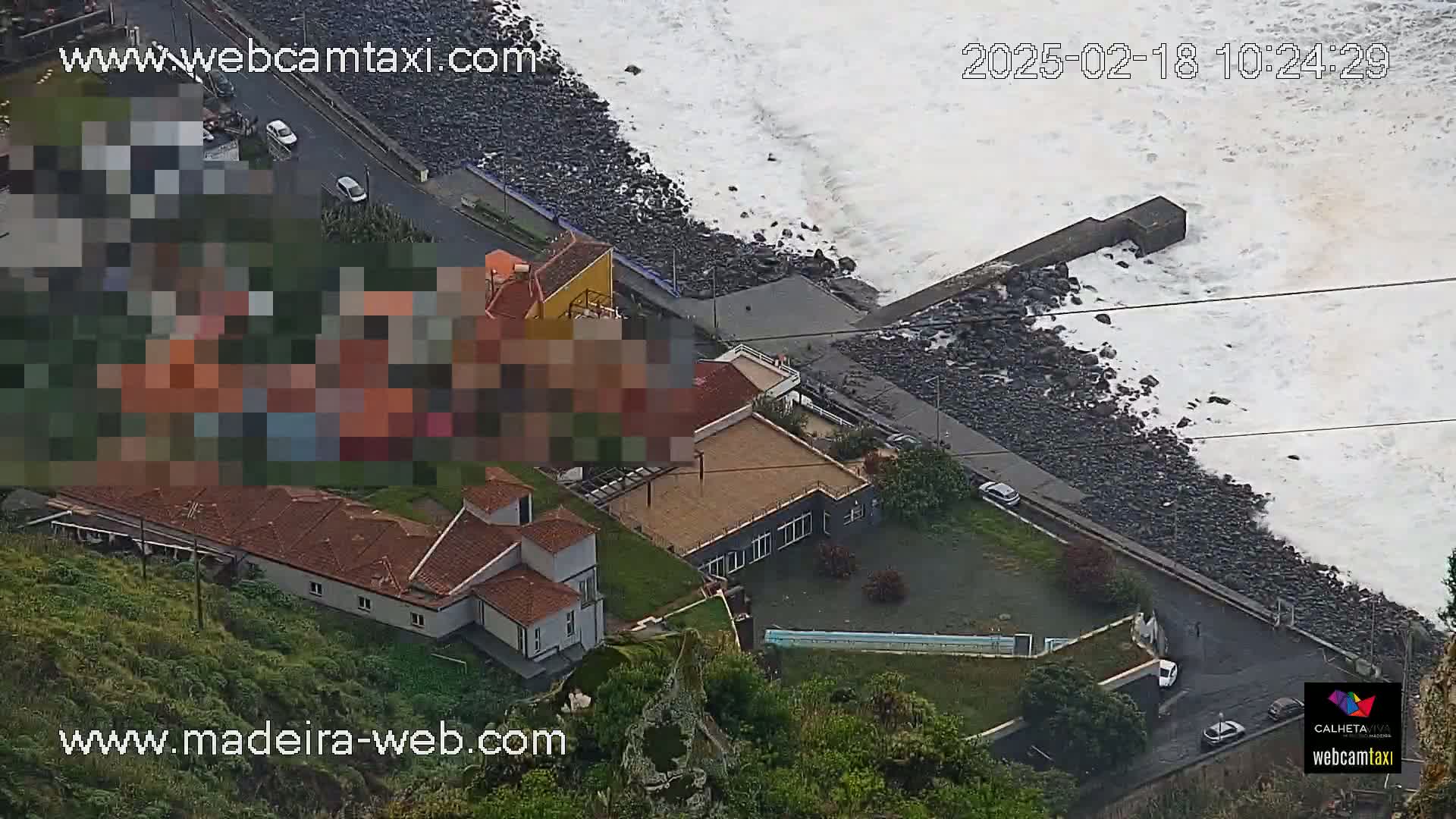 An aerial view shows a rocky coastline with crashing waves, several buildings, and a road with cars parked alongside it.