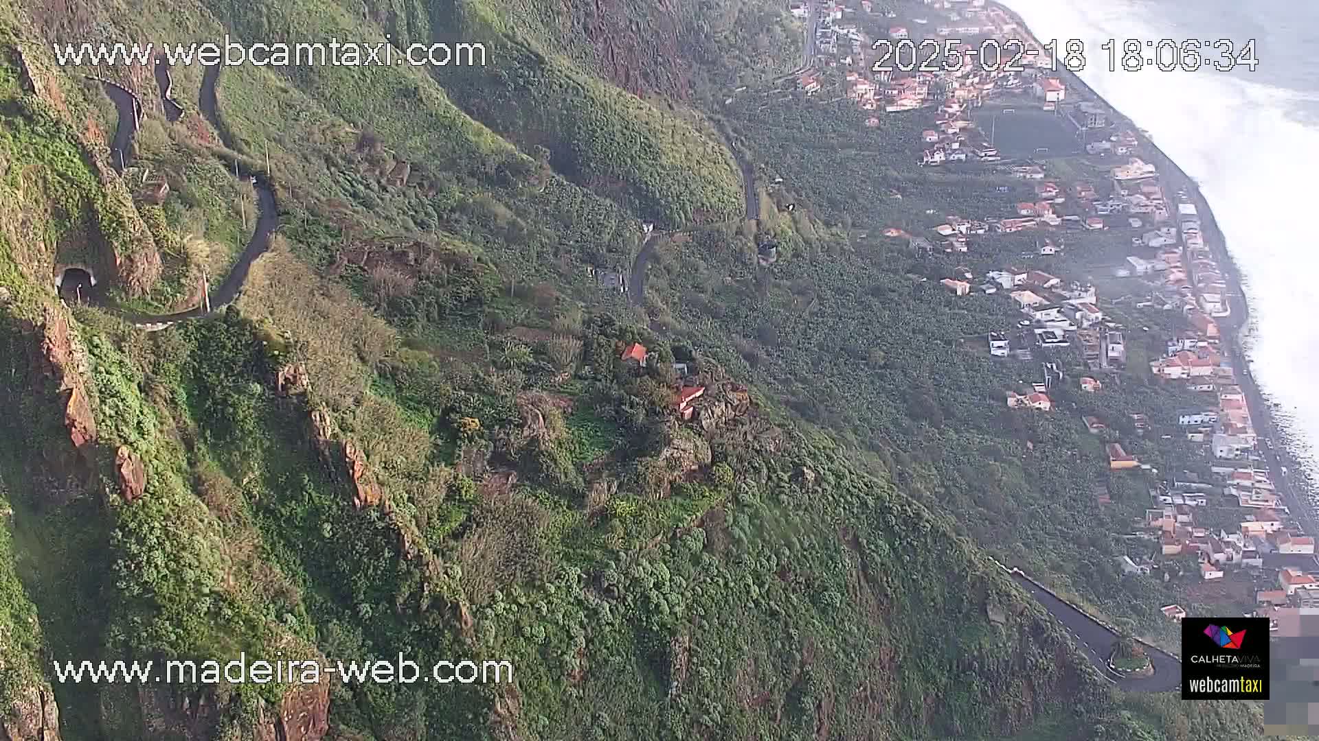 A winding road descends a lush, green hillside toward a coastal village, with the ocean visible in the background on a partly cloudy day.