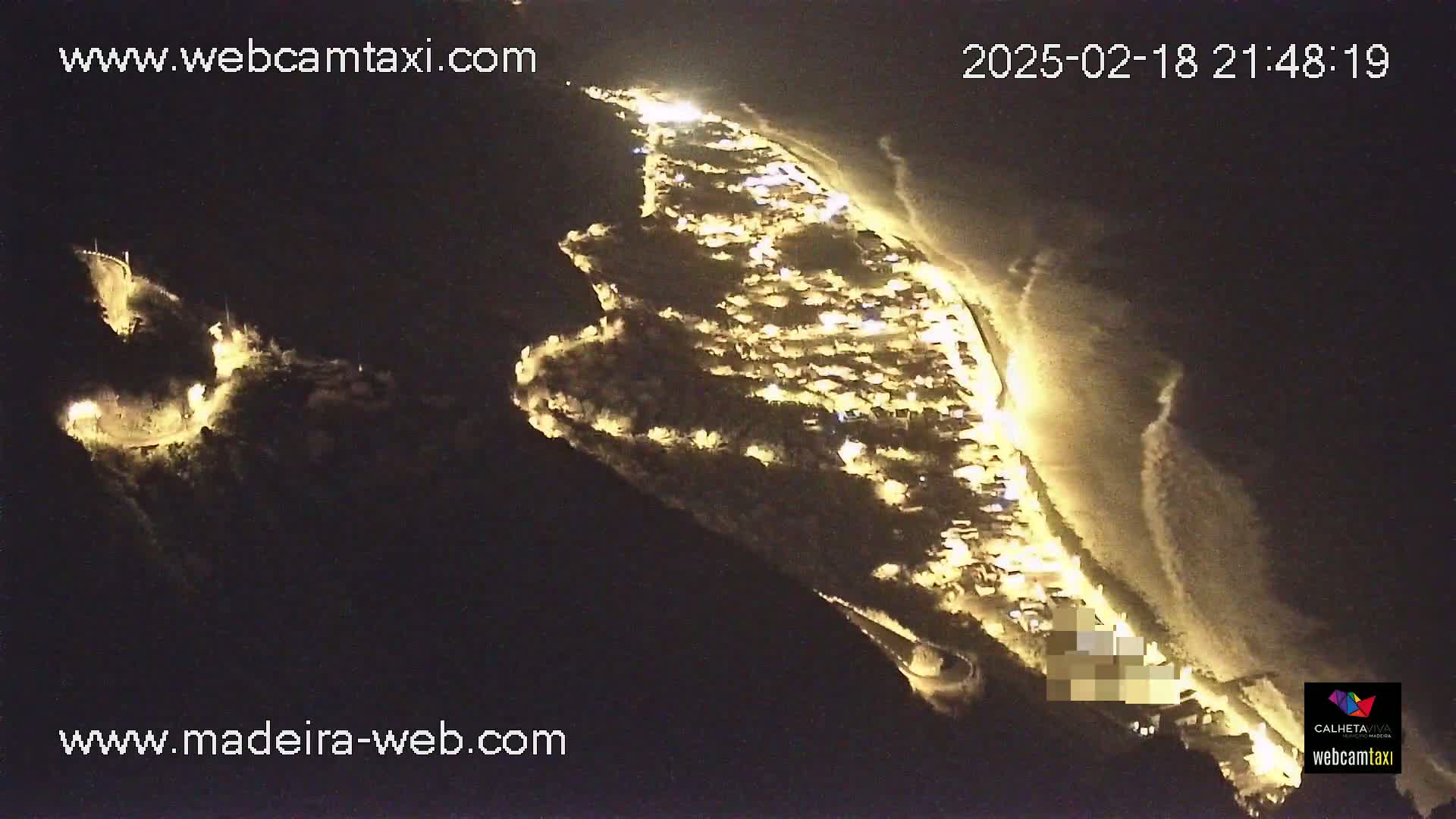 A nighttime aerial view shows a coastal town illuminated, with waves gently lapping the shore.