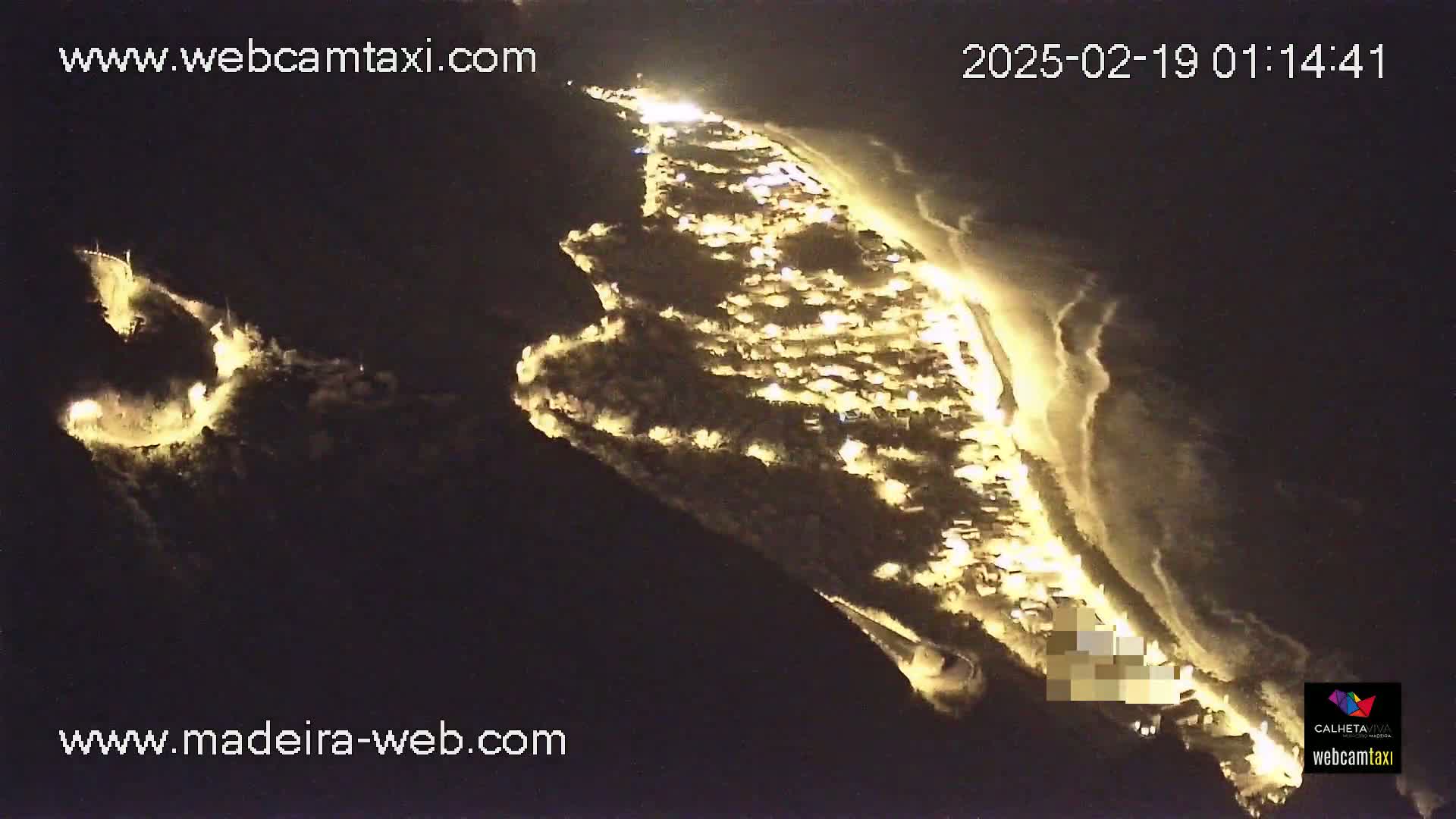 An aerial nighttime view shows a coastal town illuminated, with dark ocean waves lapping at a lit shoreline.
