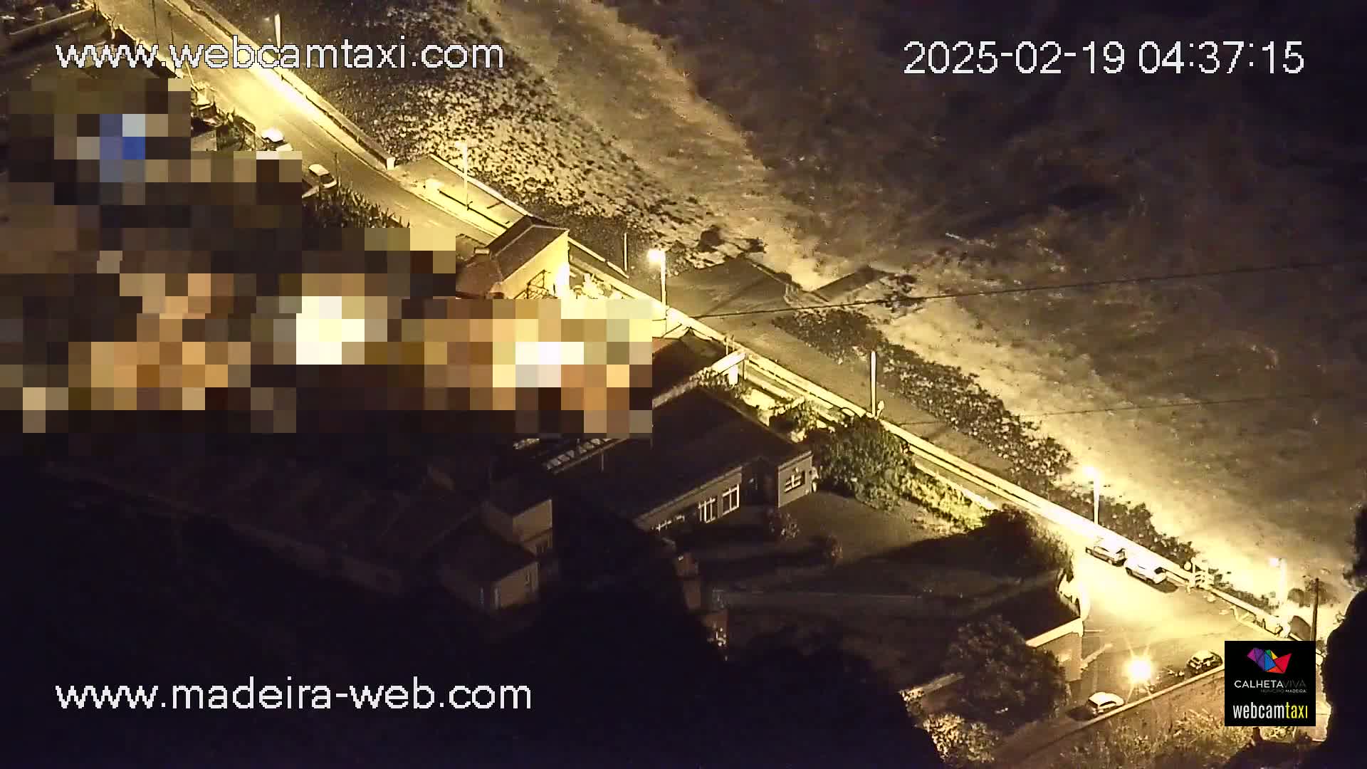 A nighttime aerial view shows a coastline with buildings and a road, illuminated by streetlights, with dark waves crashing on the shore.