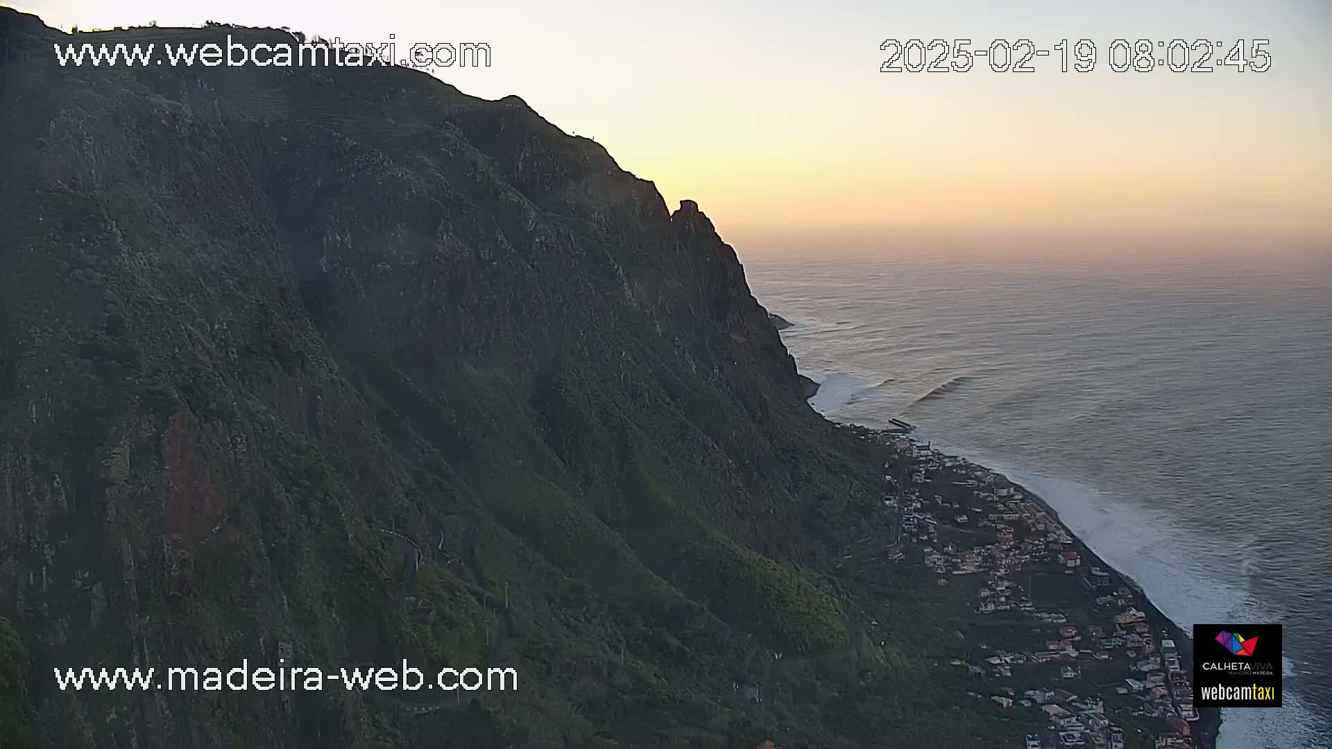 A high-angle view shows a steep, green cliffside descending to a dark-sand beach and ocean at sunset.