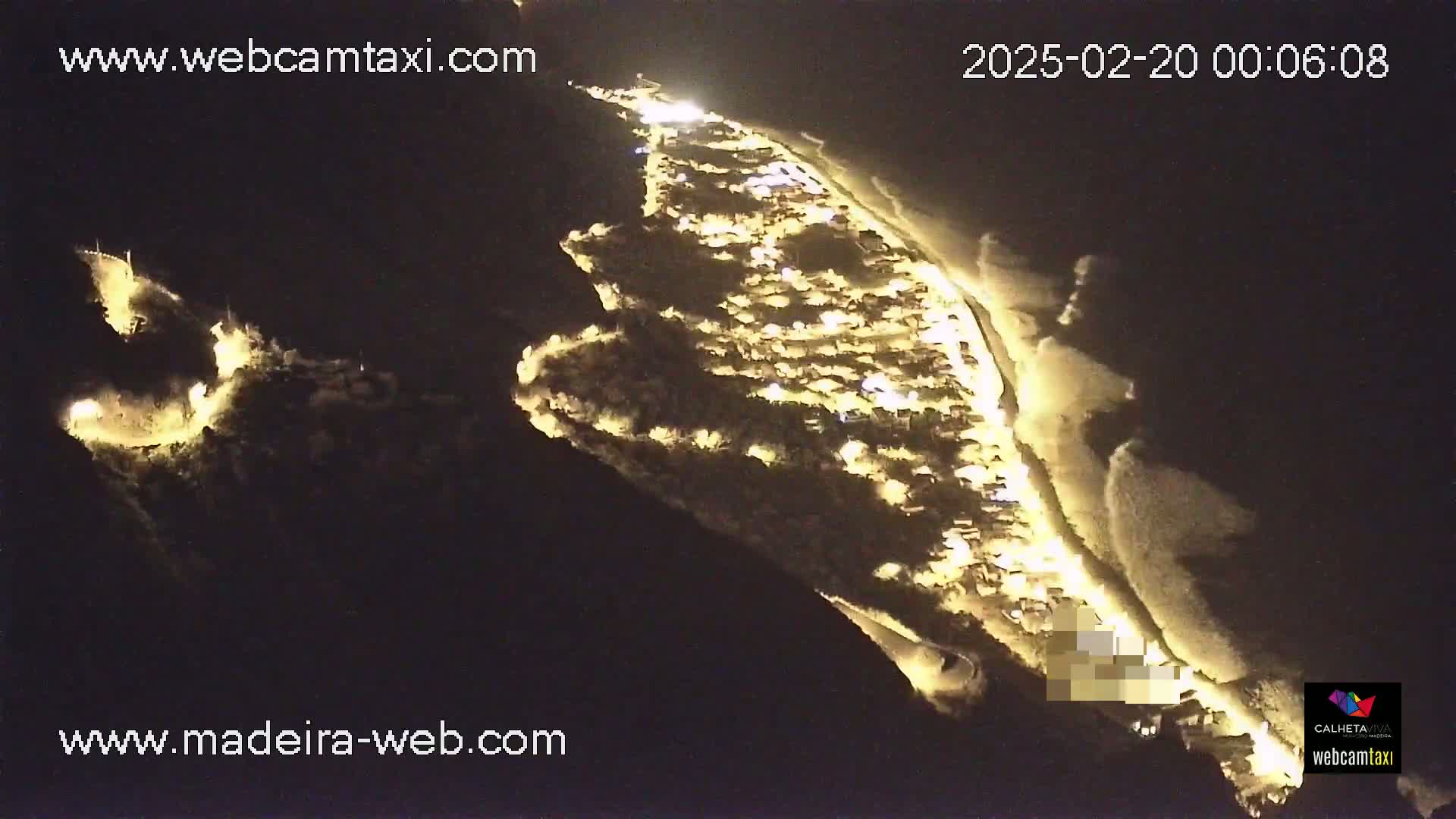 A nighttime aerial view shows a coastal town illuminated, with waves gently breaking on the shore.