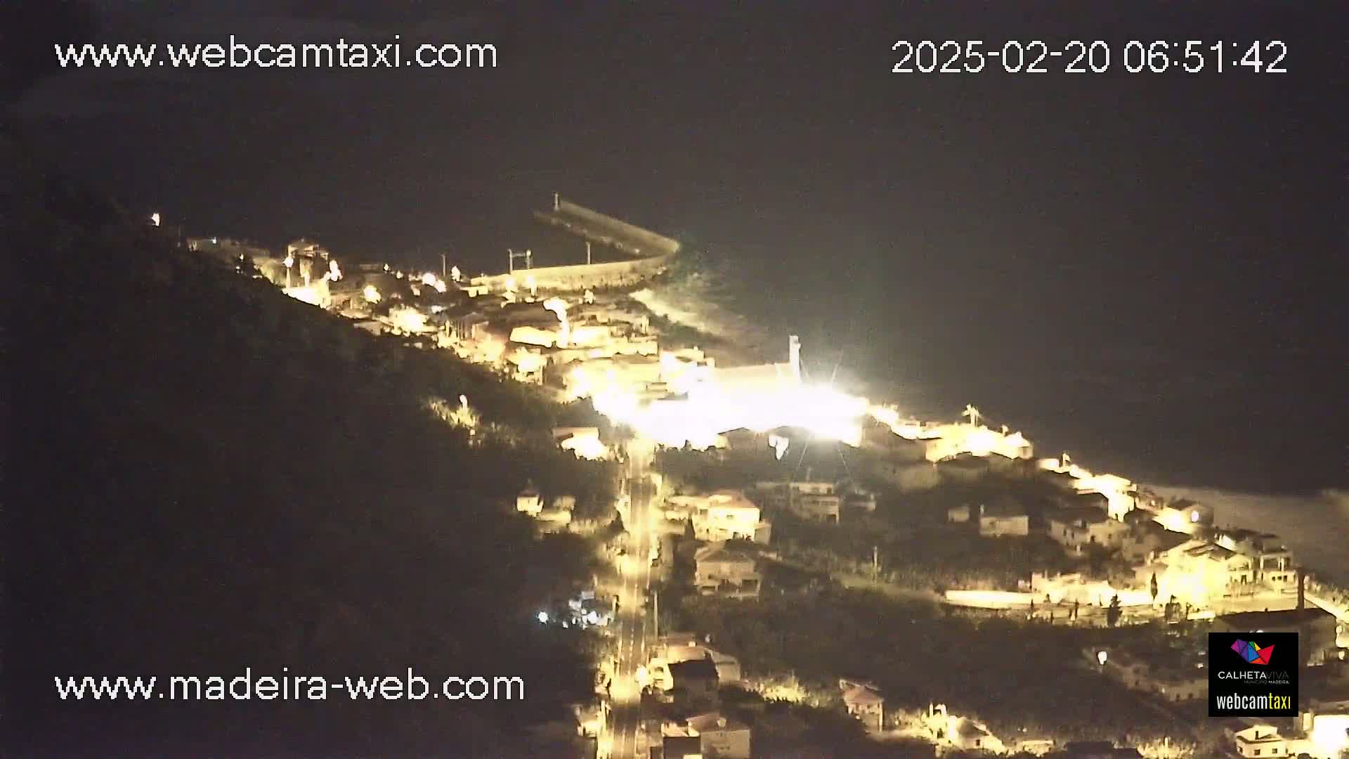 A nighttime aerial view shows a small, illuminated town nestled on a hillside overlooking a dark ocean.