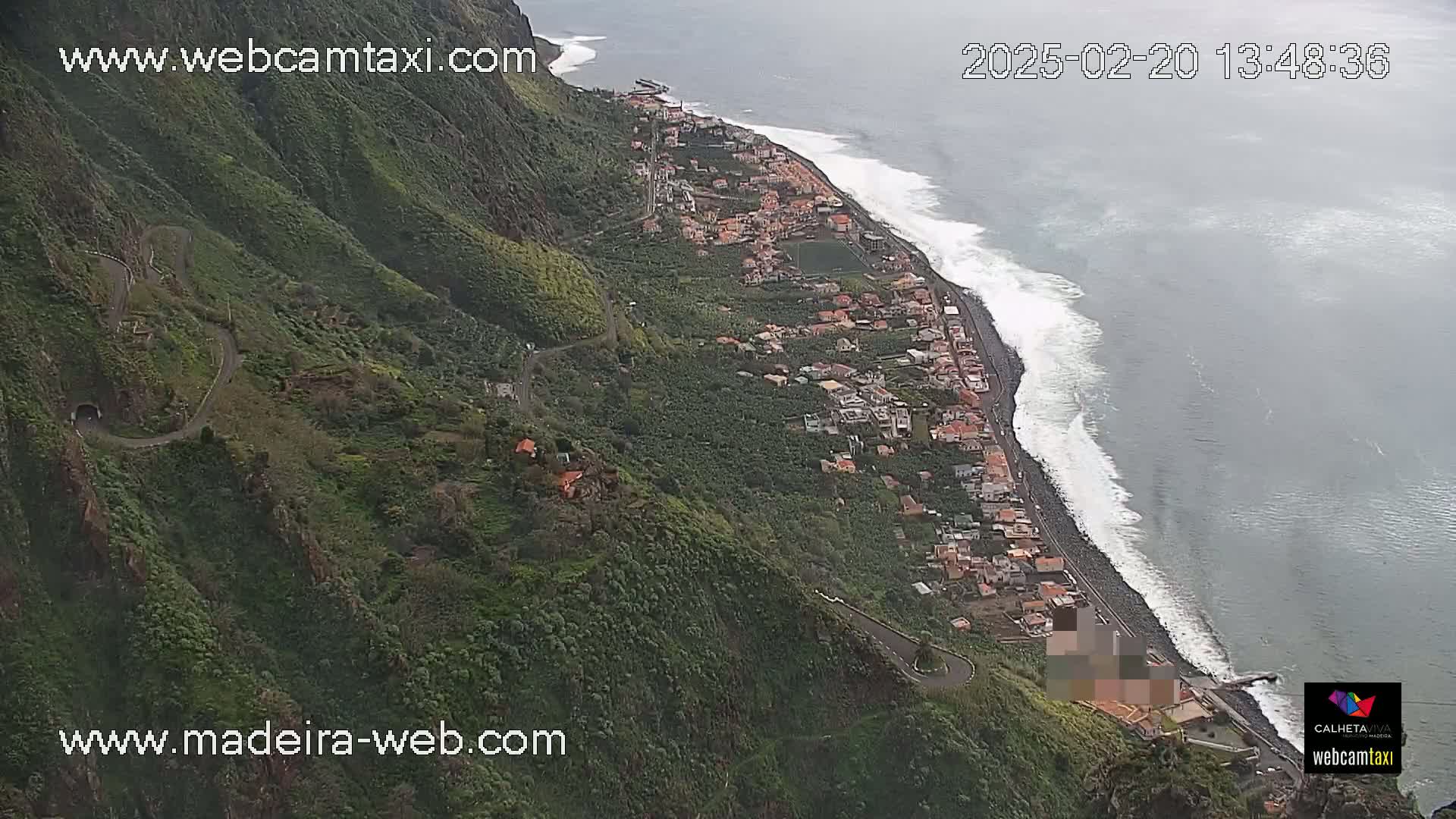An aerial view shows a coastal town nestled between a verdant mountainside and a dark-blue ocean with white-capped waves on an overcast day.