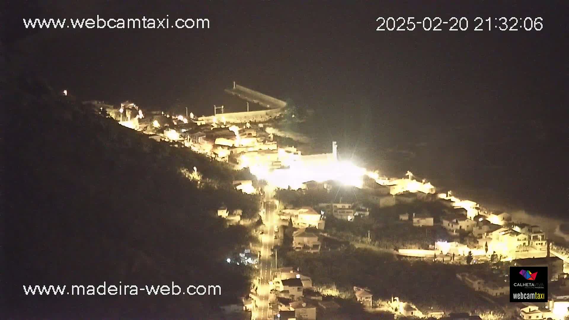 A nighttime, aerial view of a coastal town illuminated by streetlights, situated on a hillside overlooking a dark body of water.