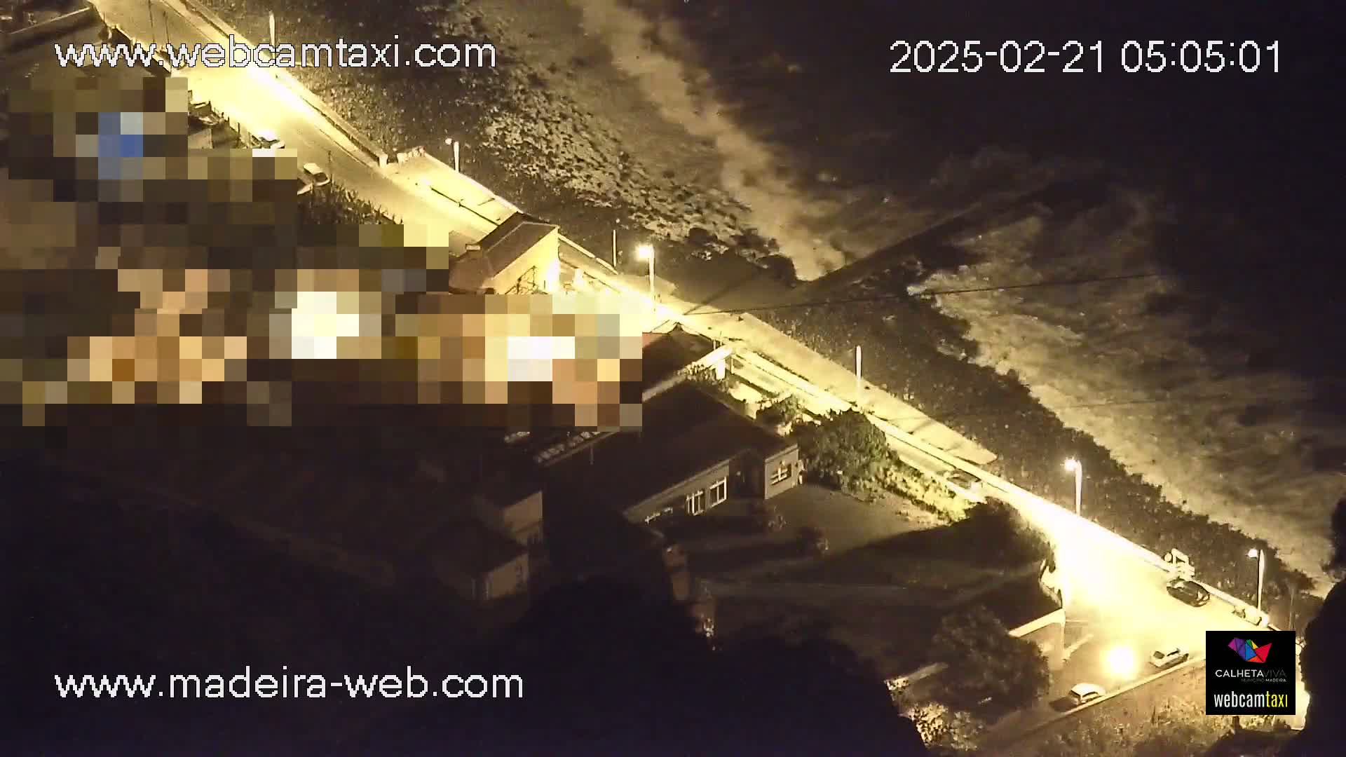 An aerial nighttime view shows a coastal road with parked cars, buildings, and large waves crashing on the shore.