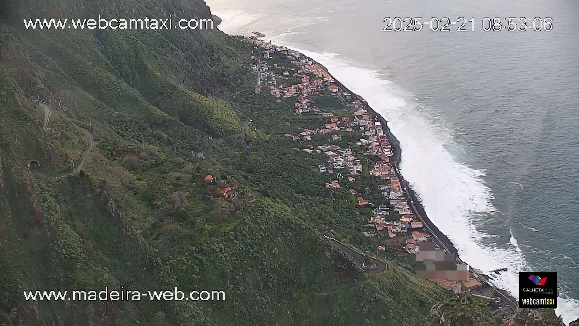 An aerial view shows a coastal town nestled on a steep, verdant hillside overlooking a dark-blue ocean with white-capped waves.
