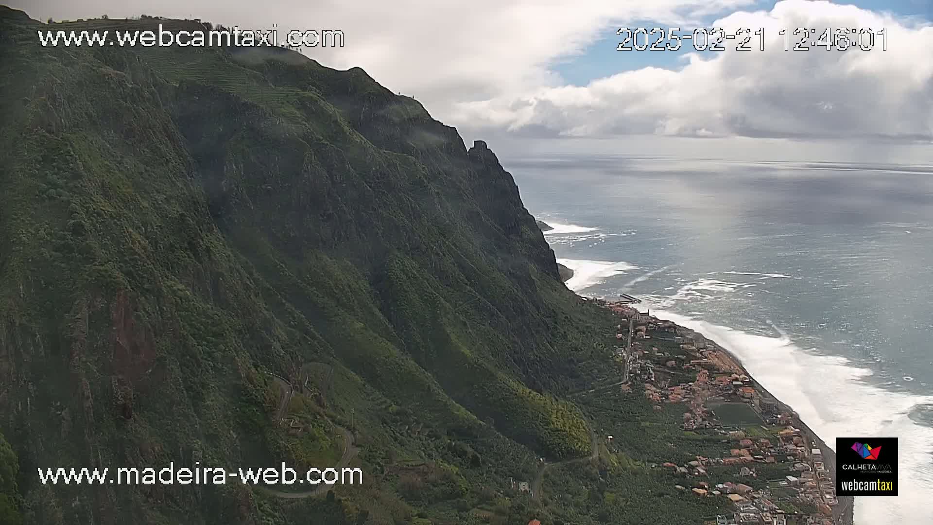 A verdant, steep cliffside descends to a coastal town, with a partly cloudy sky and ocean waves breaking along the shore.