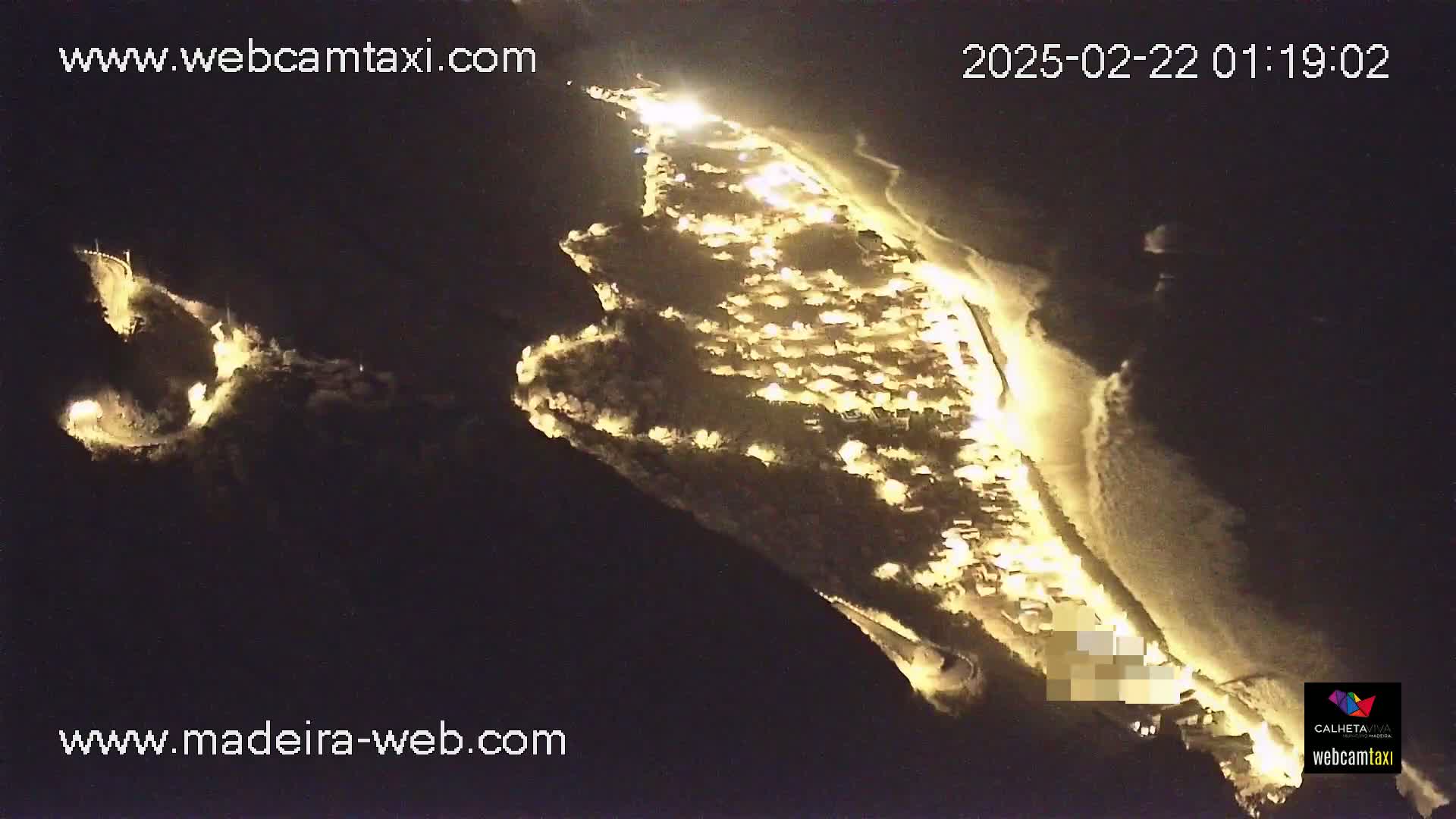 A nighttime aerial view shows a coastal town illuminated, with waves gently lapping at a sandy shore.