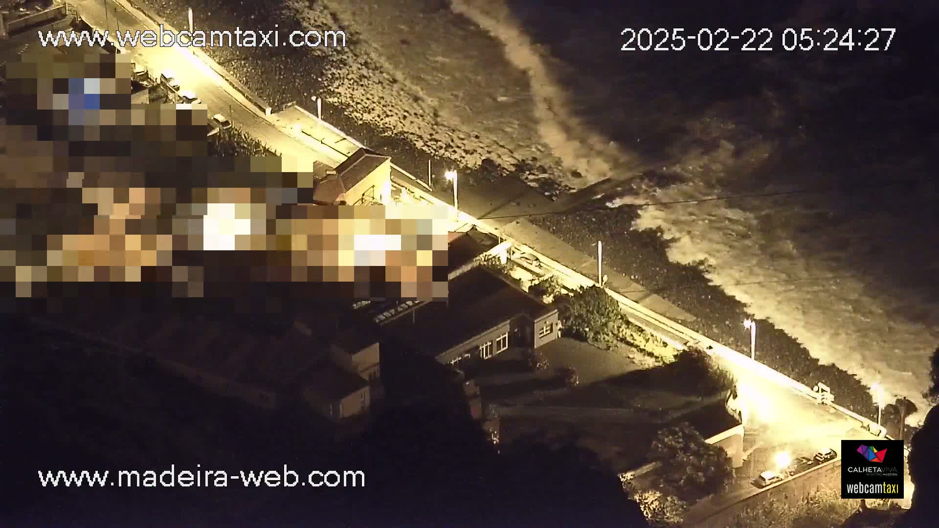 An aerial nighttime view shows large waves crashing against a seawall next to buildings and a road.