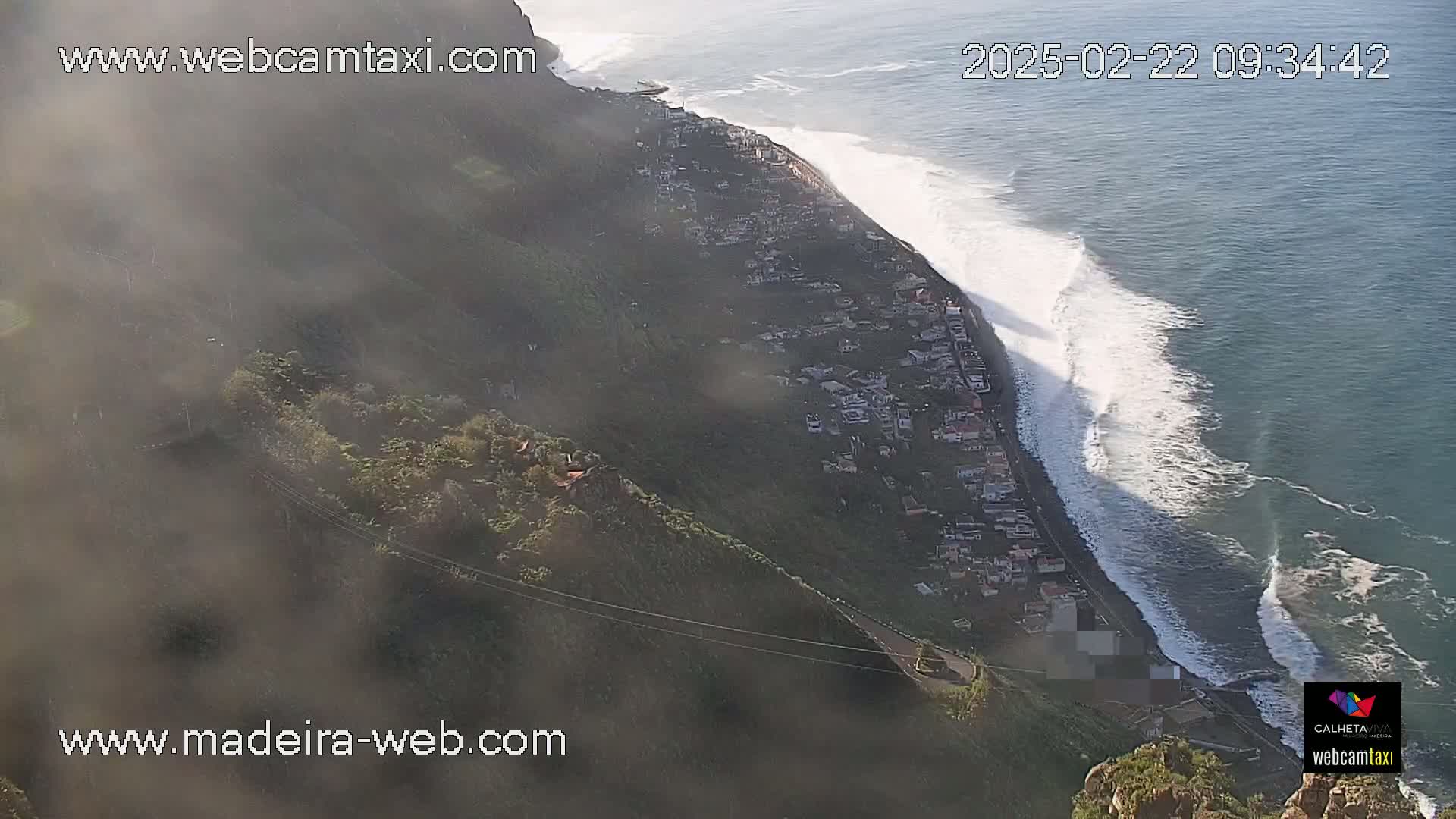 A coastal town nestled on a steep hillside overlooking a dark-blue ocean with large, white-capped waves, partially obscured by morning mist.