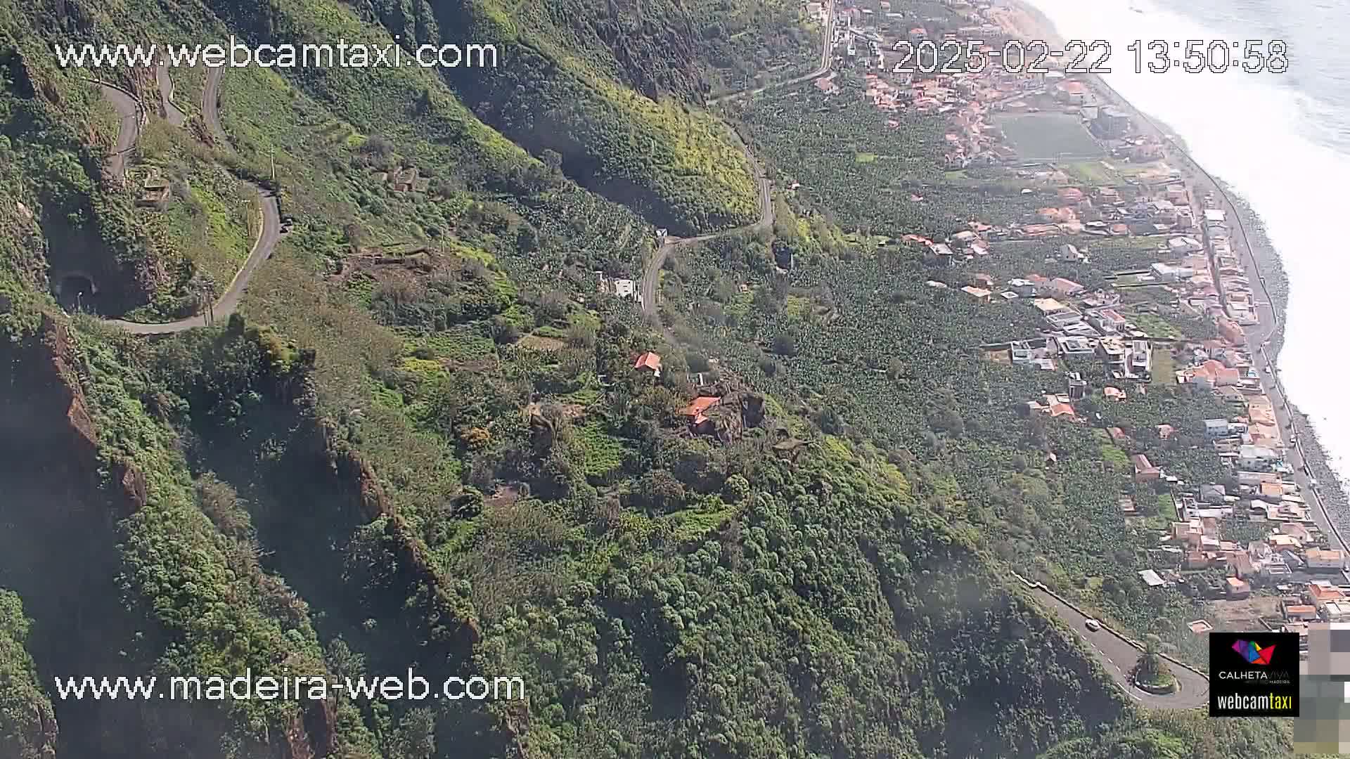 A winding road snakes down a lush, green hillside toward a coastal town and ocean under sunny skies.
