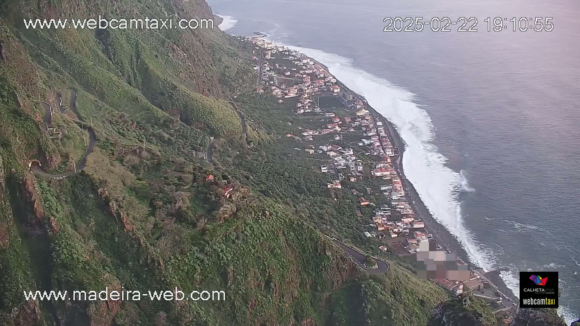 A coastal town nestled along a rocky cliffside, with a winding road curving down the mountain and large waves crashing against a dark-colored beach under a mostly cloudy sky.
