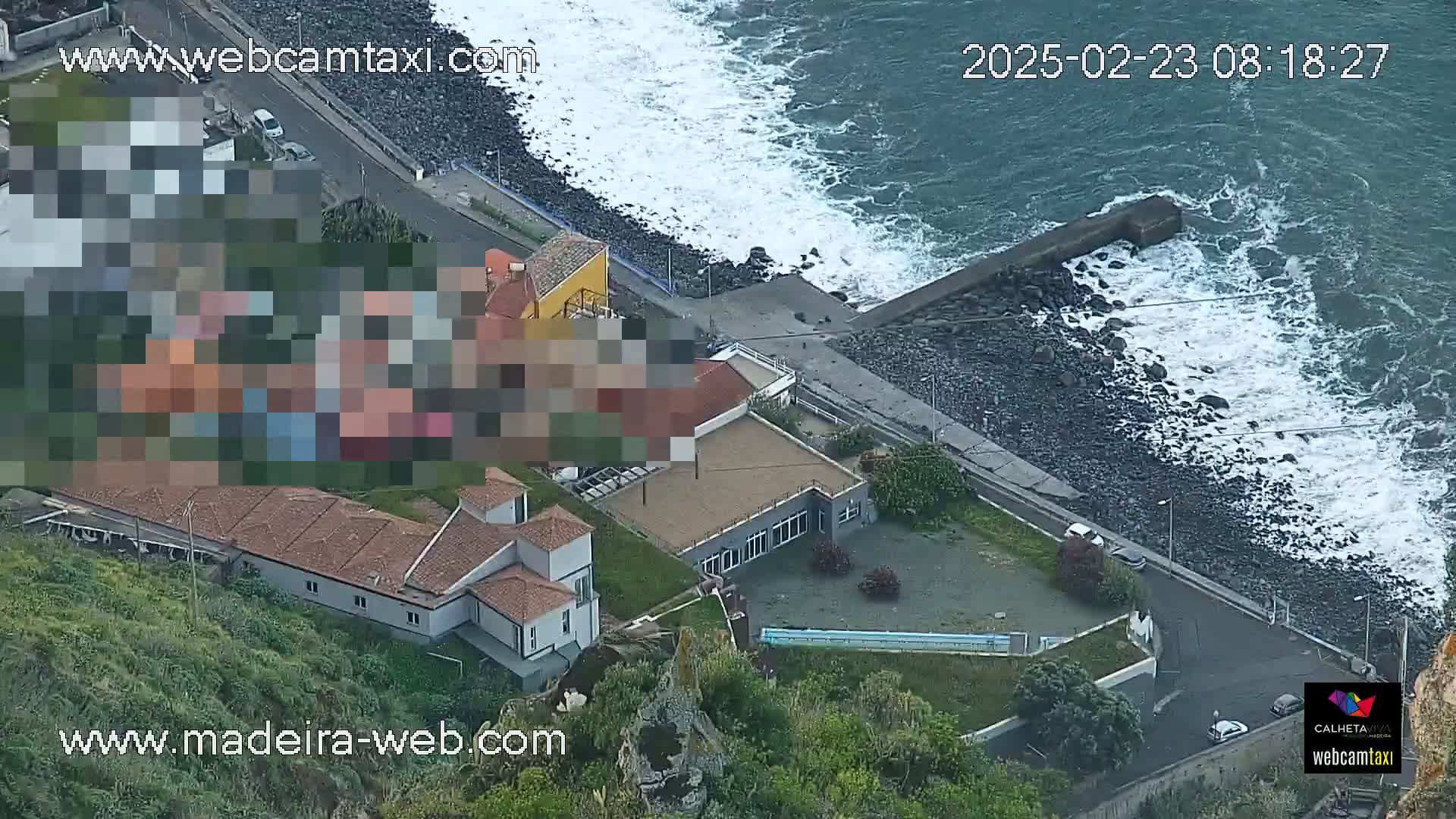 An aerial view shows a rocky coastline with ocean waves crashing against a seawall near buildings, some of which are partially obscured.