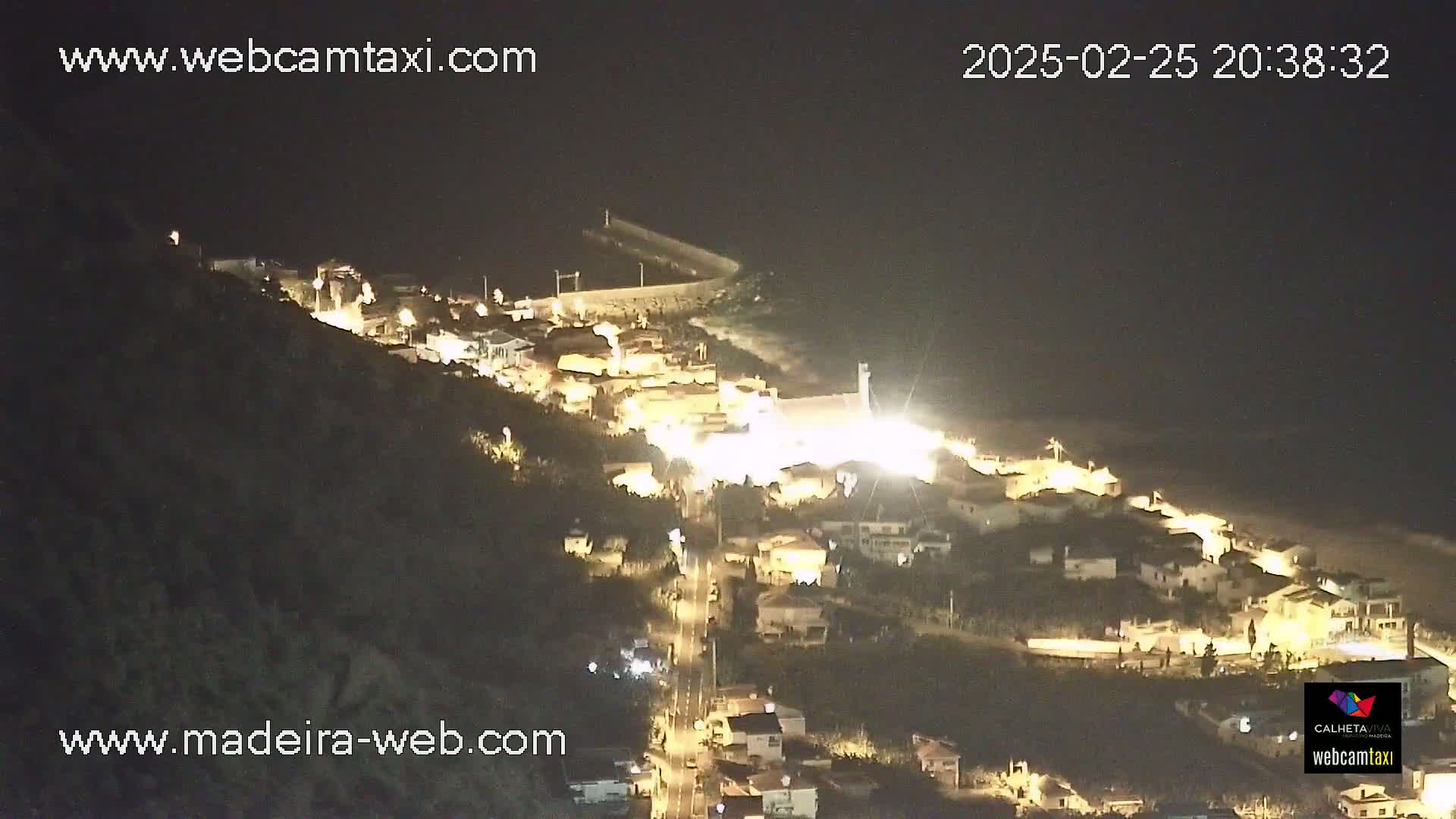 A nighttime, aerial view shows a coastal town illuminated, nestled between a dark hillside and a calm ocean.