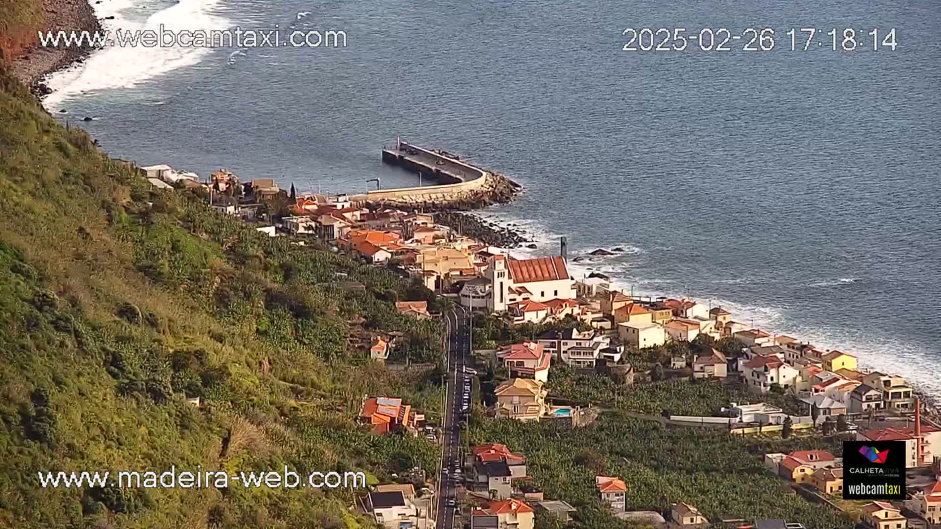 A coastal town with red-roofed houses clings to a hillside overlooking a calm ocean with gentle waves, under a clear sky.