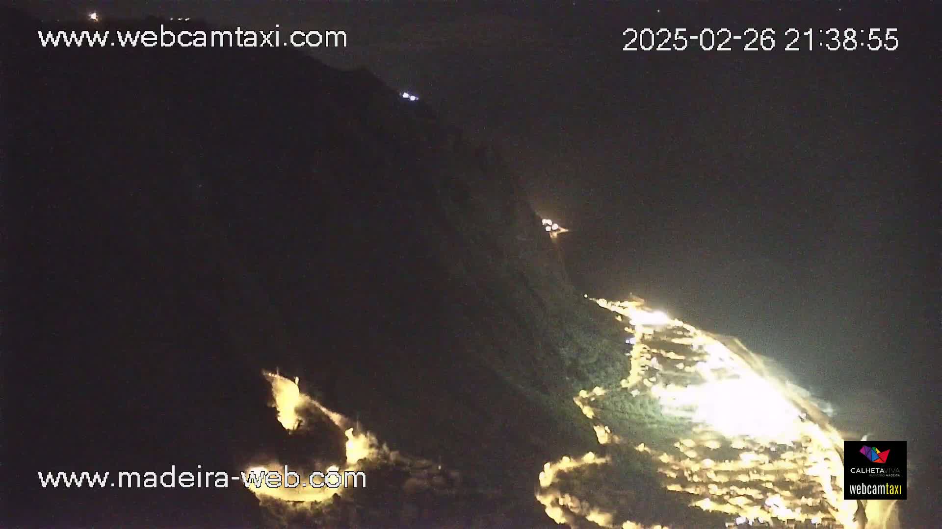 A nighttime long shot of a coastal area with illuminated roads and buildings on a dark hillside.
