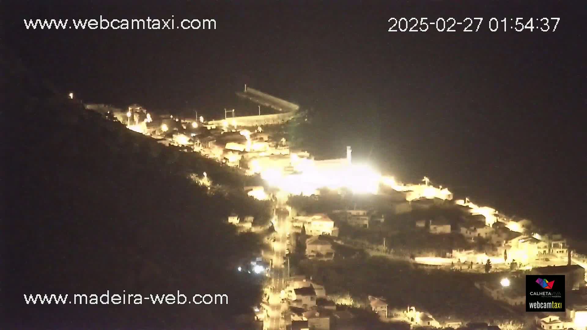 A nighttime aerial view shows a brightly lit village nestled on a hillside, with a dark body of water visible in the distance under a clear, dark sky.