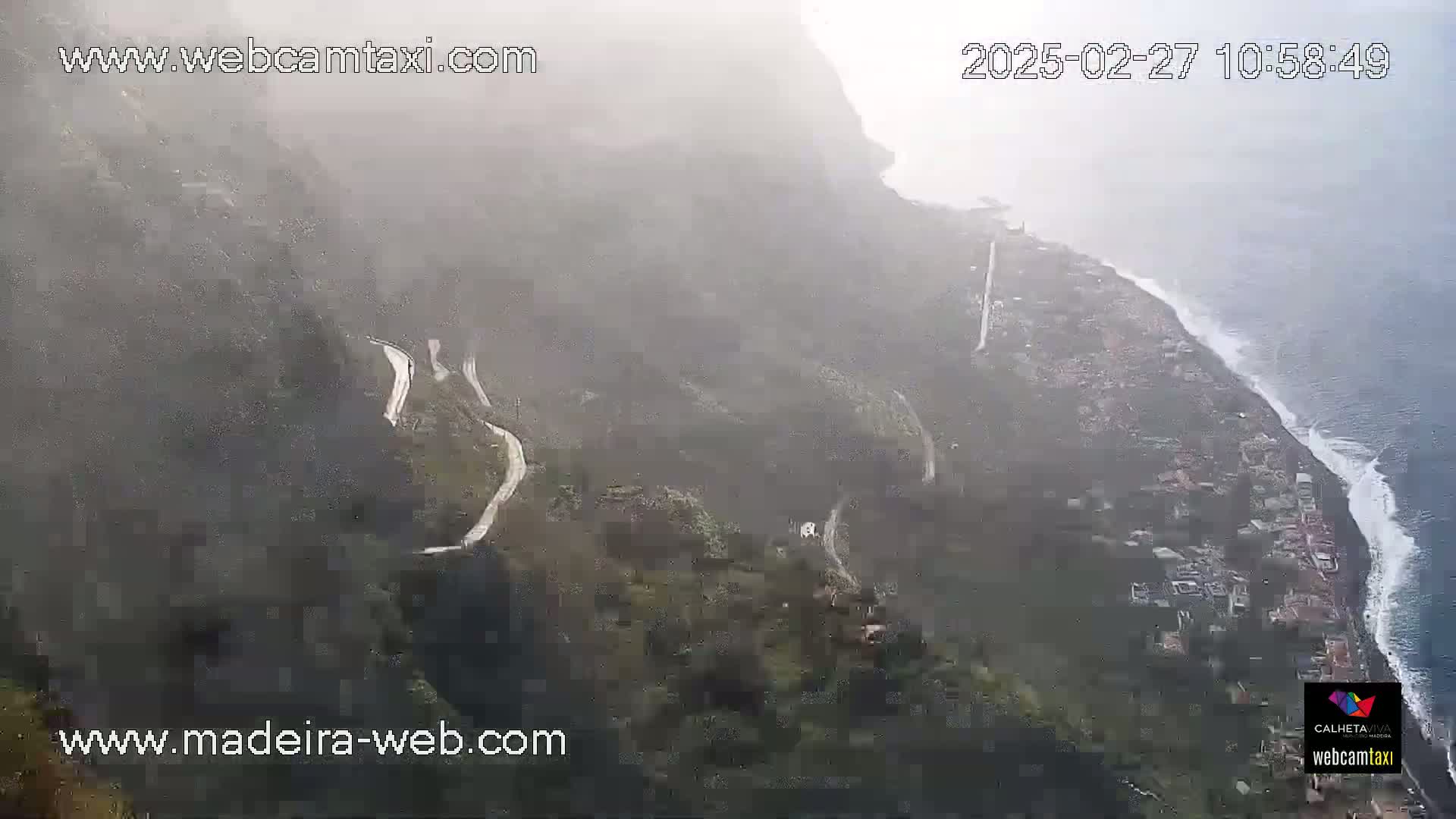 A misty, high-angle view shows a winding road snaking down a steep, green hillside toward a coastal town and ocean with breaking waves.