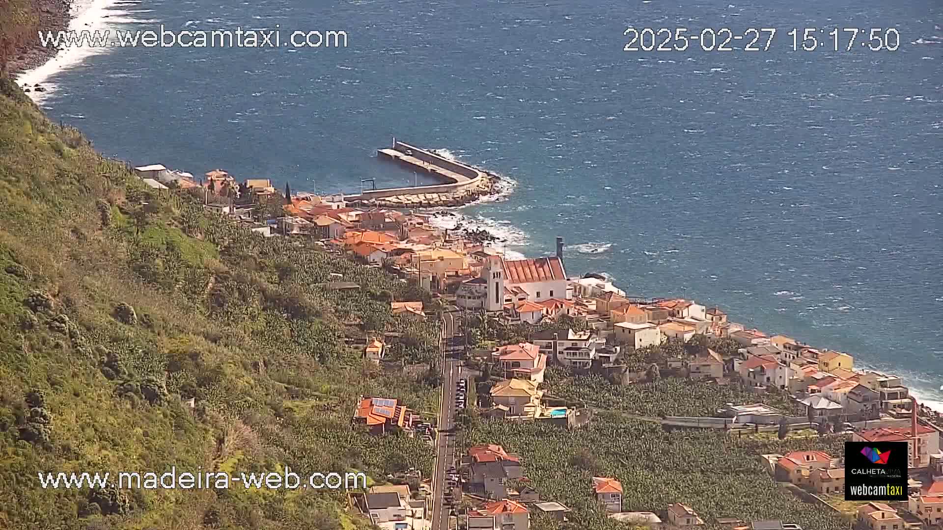 A coastal town with red-roofed buildings and banana plantations on a hillside overlooking a wavy ocean under a sunny sky.