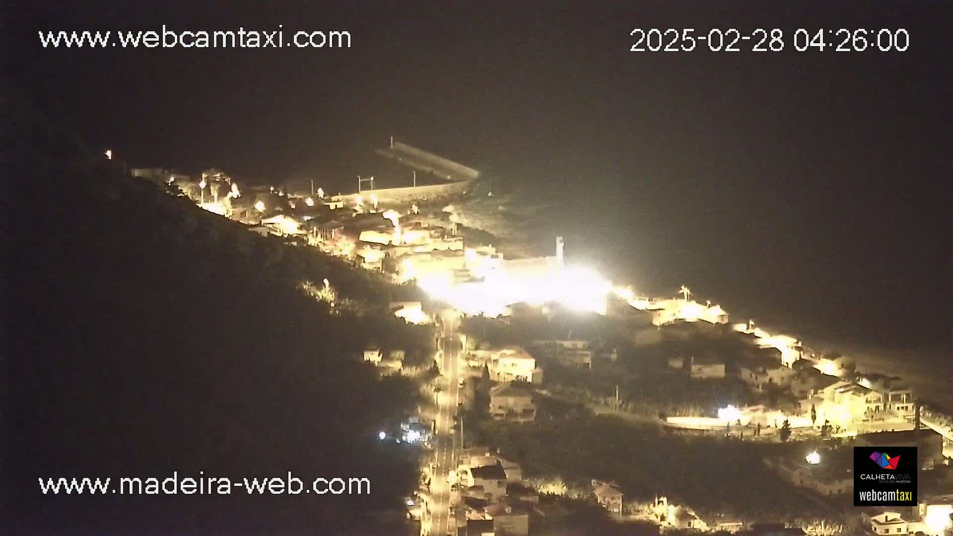 A nighttime, aerial view of a coastal town illuminated by streetlights, nestled on a hillside overlooking the ocean.