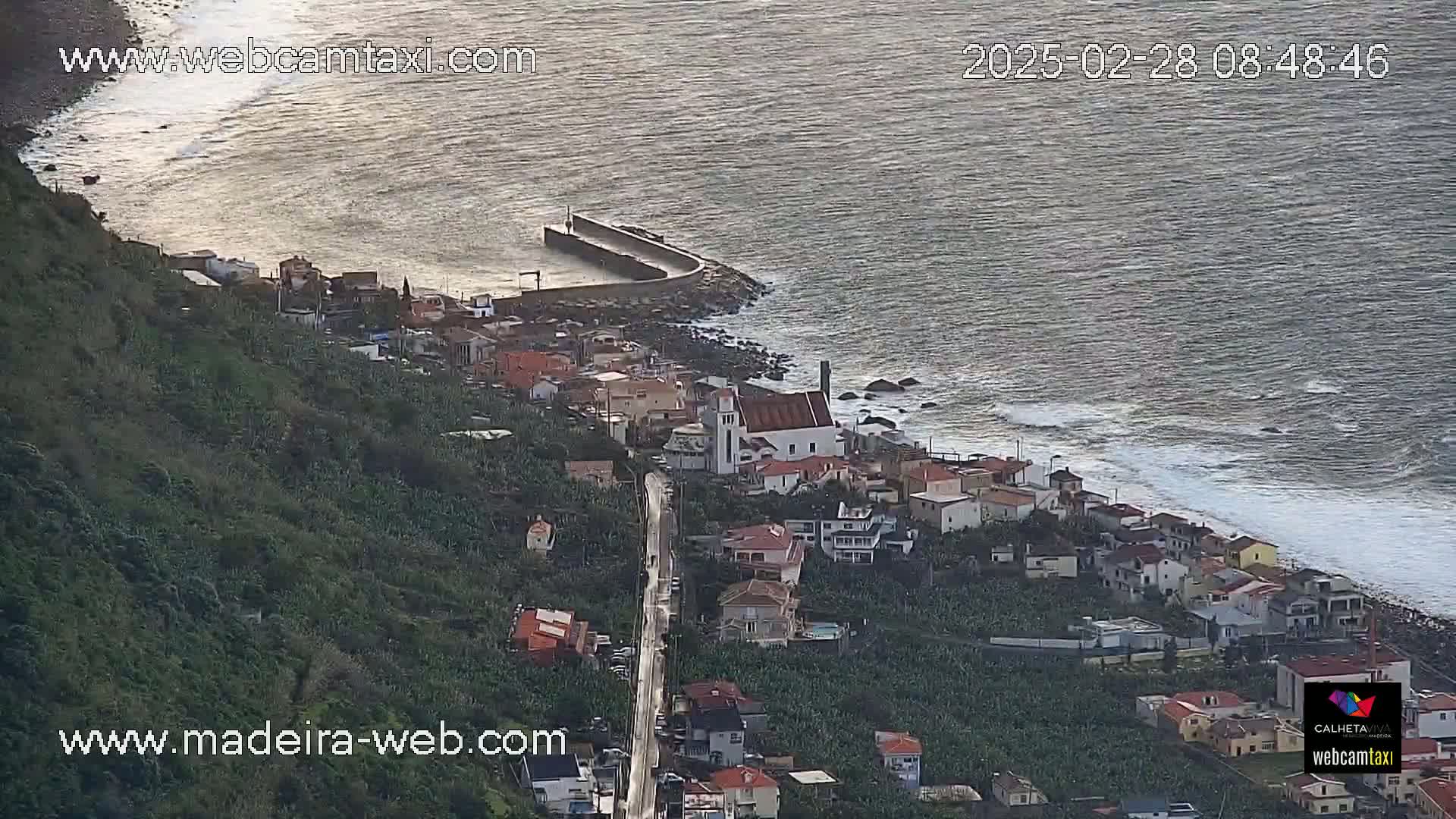 A coastal town with a church nestled between a lush green hillside and a rocky shoreline with moderate waves under a partly cloudy sky.